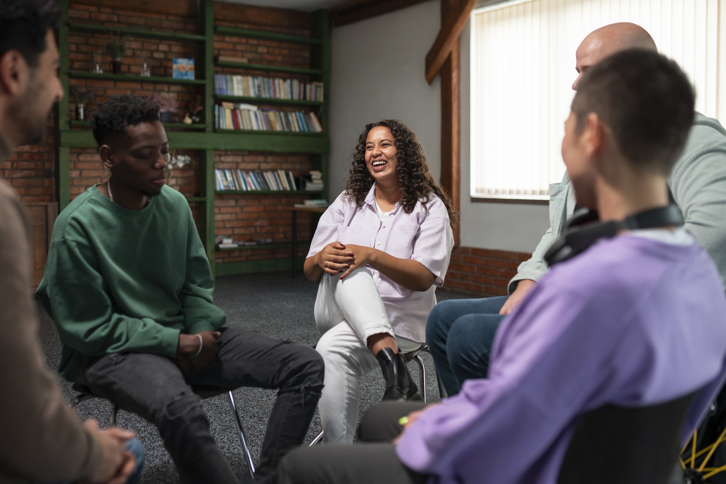 A diverse group of people sitting in a circle in a room with bookshelves, engaged in a group discussion with a woman laughing in the center.