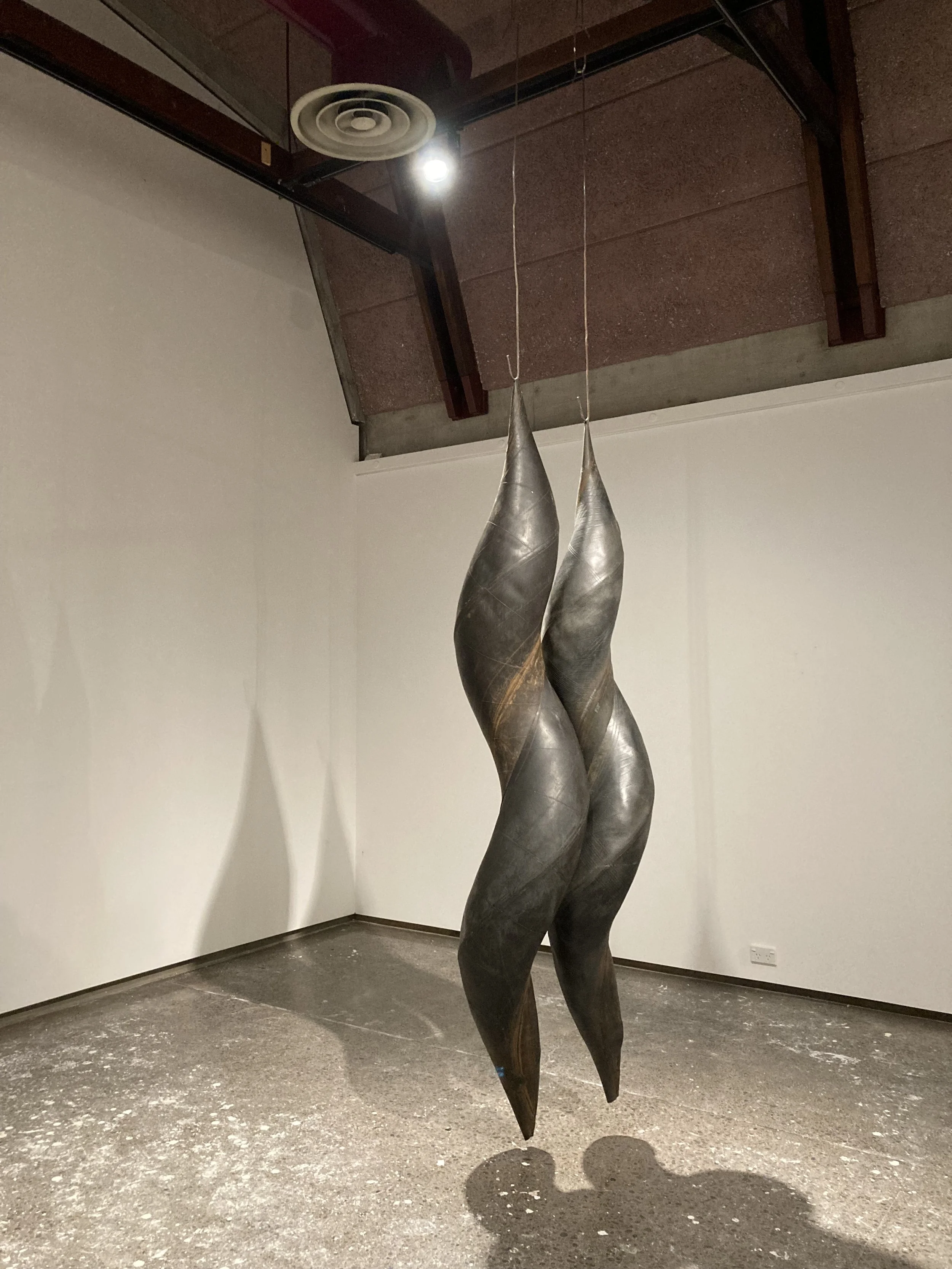 Sculpture of two elongated, twisted, and polished metal figures hanging from the ceiling in an art gallery, casting shadows on the ground.