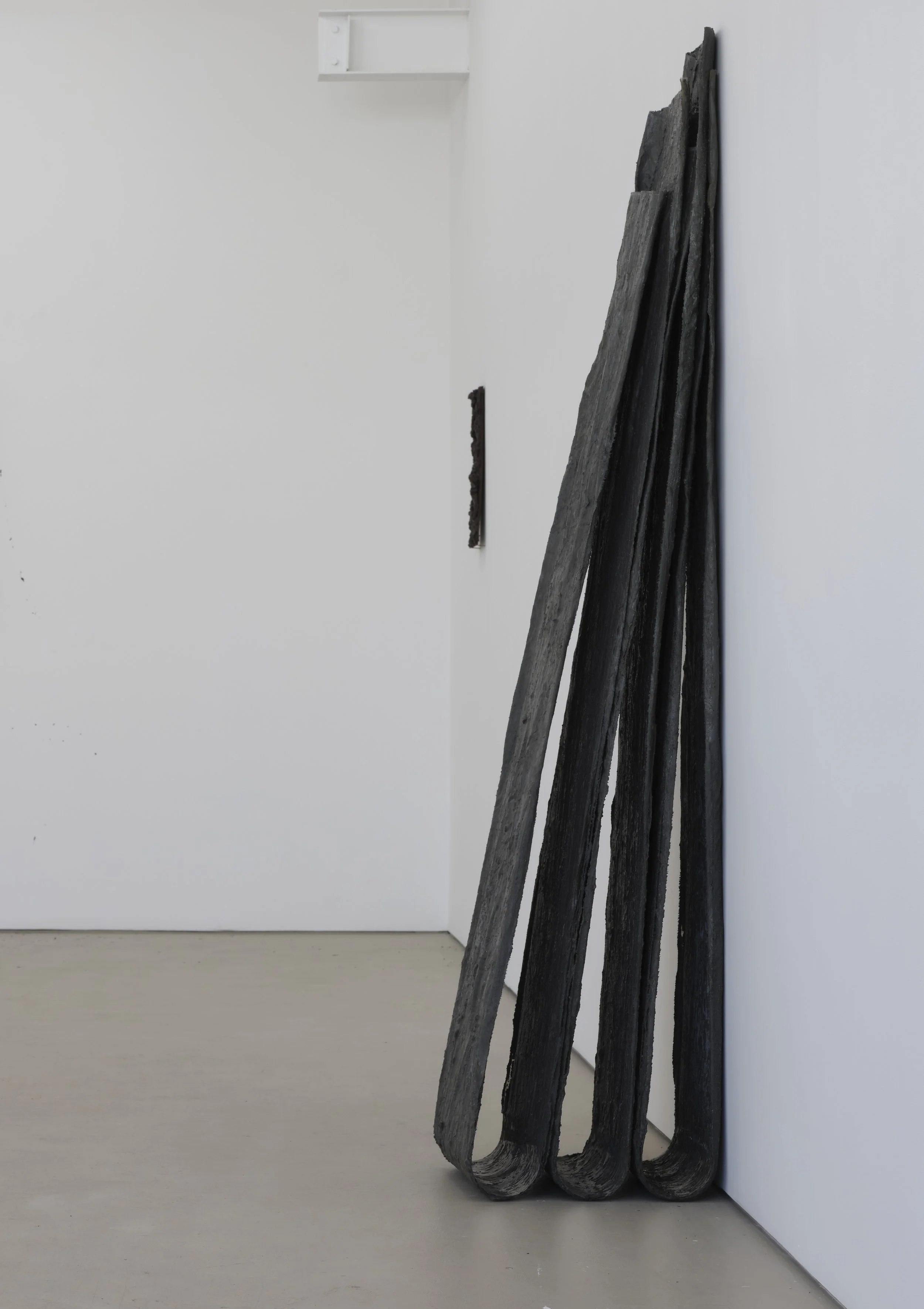 Art installation of four large, weathered, dark wooden planks leaning against a white gallery wall in a minimalist art gallery space.