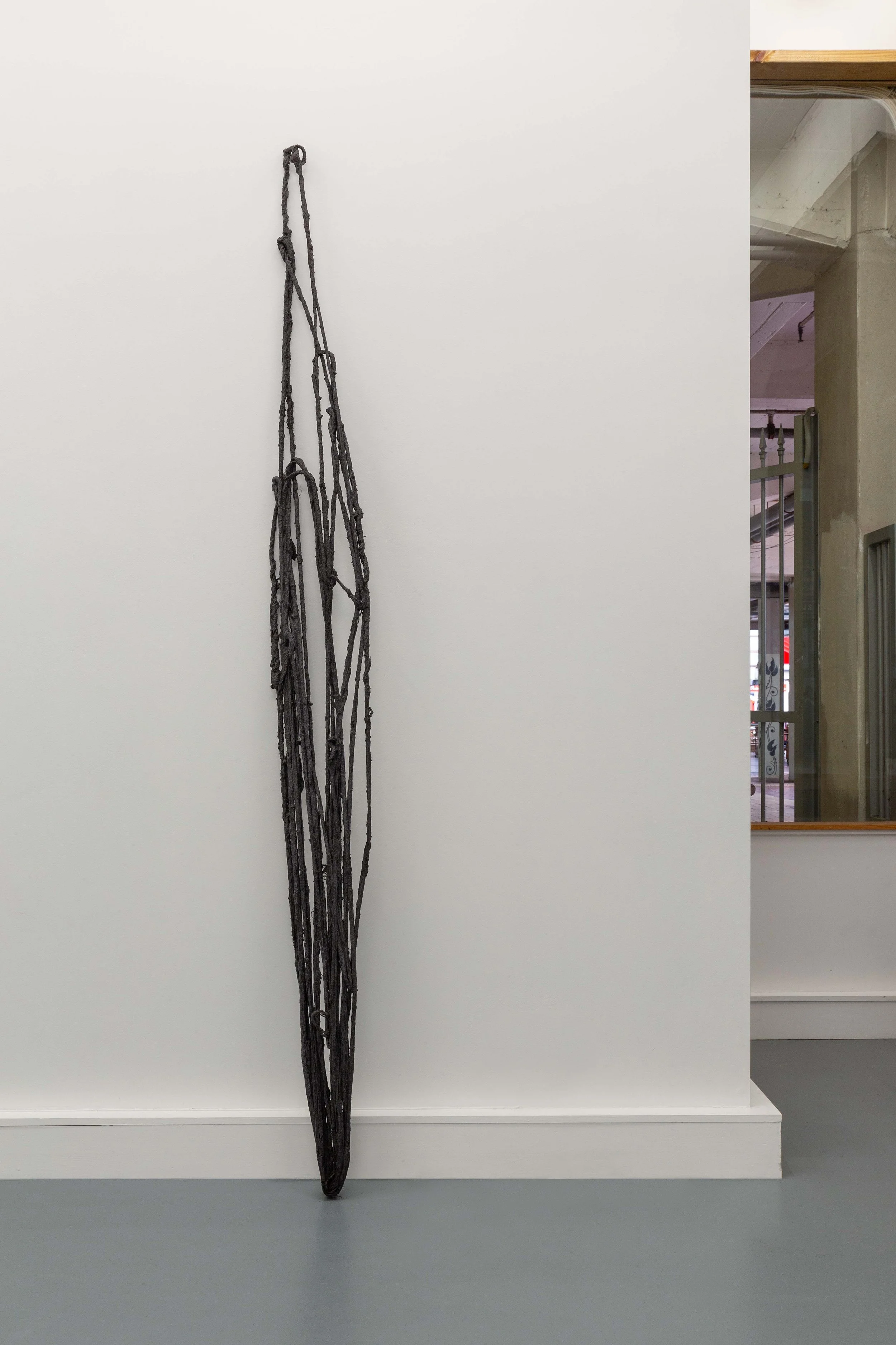 A tall, black, abstract sculpture made of intertwined, carved wooden sticks leaning against a white gallery wall.