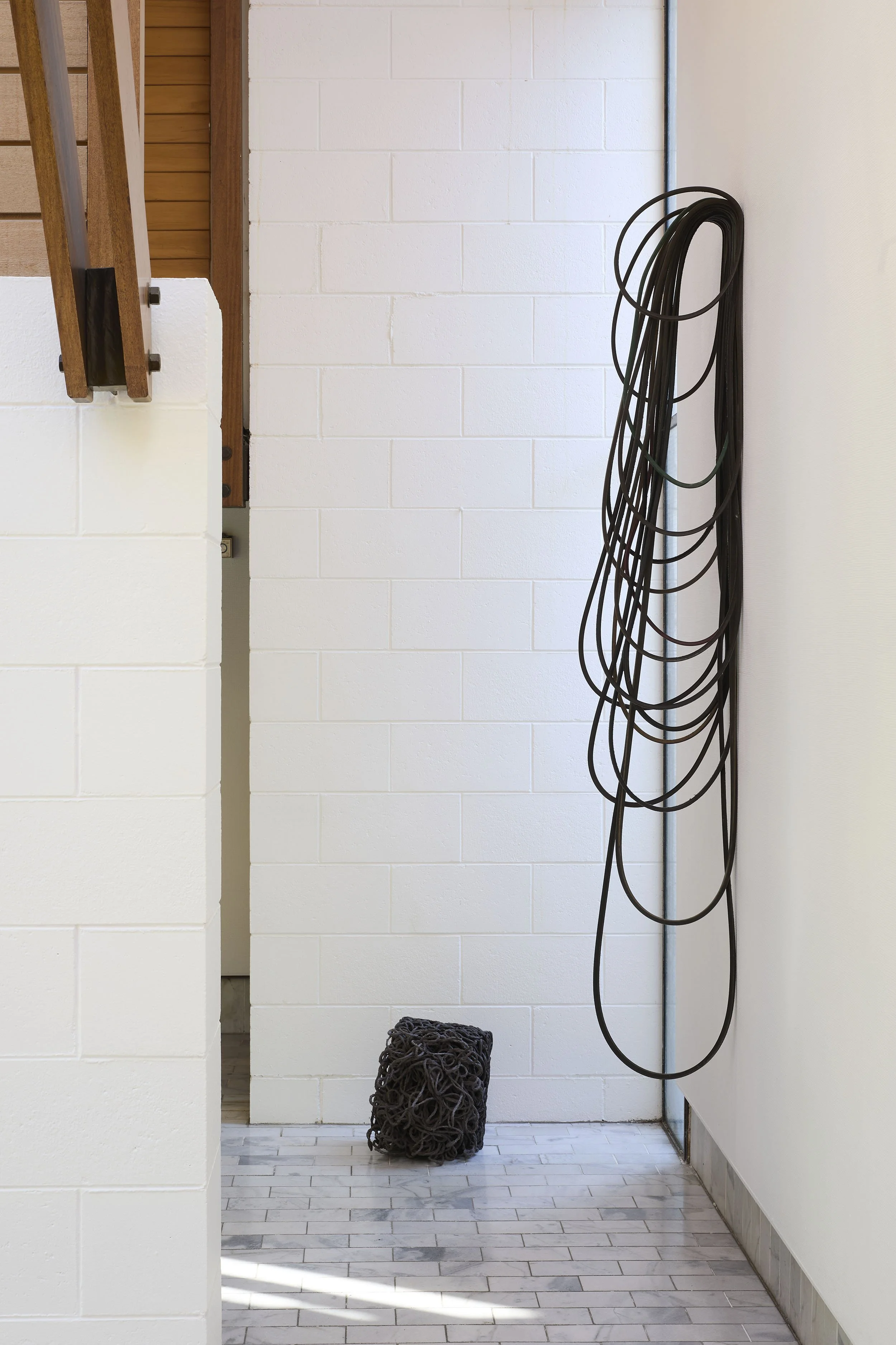 Indoor space with white brick walls, a wooden staircase, black tangled cables or hoses hanging on the wall, and a black cube-shaped sculpture on the marble floor.