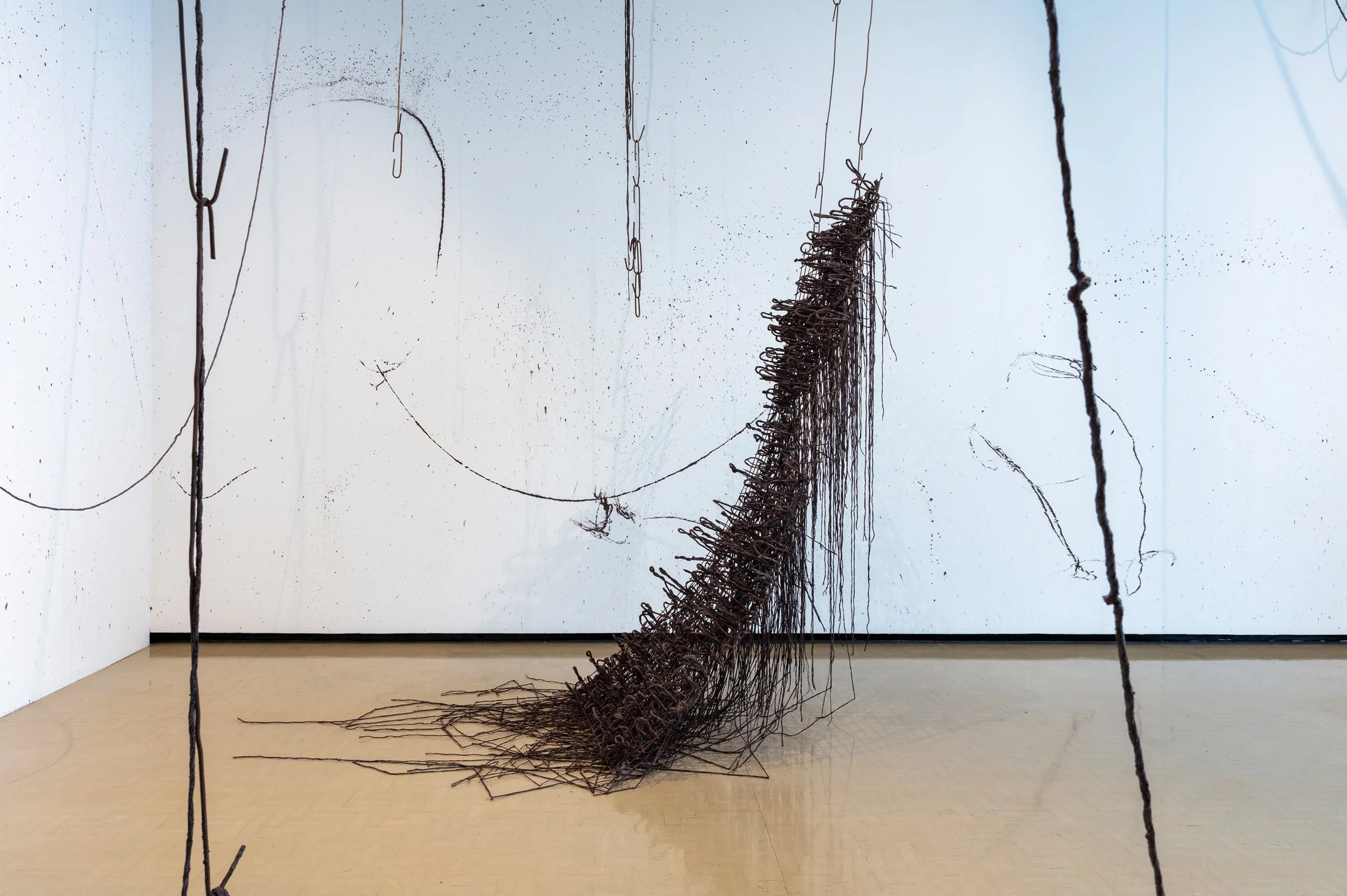 Abstract sculpture made of tangled black wire cascading from the ceiling to the floor, displayed in a gallery.