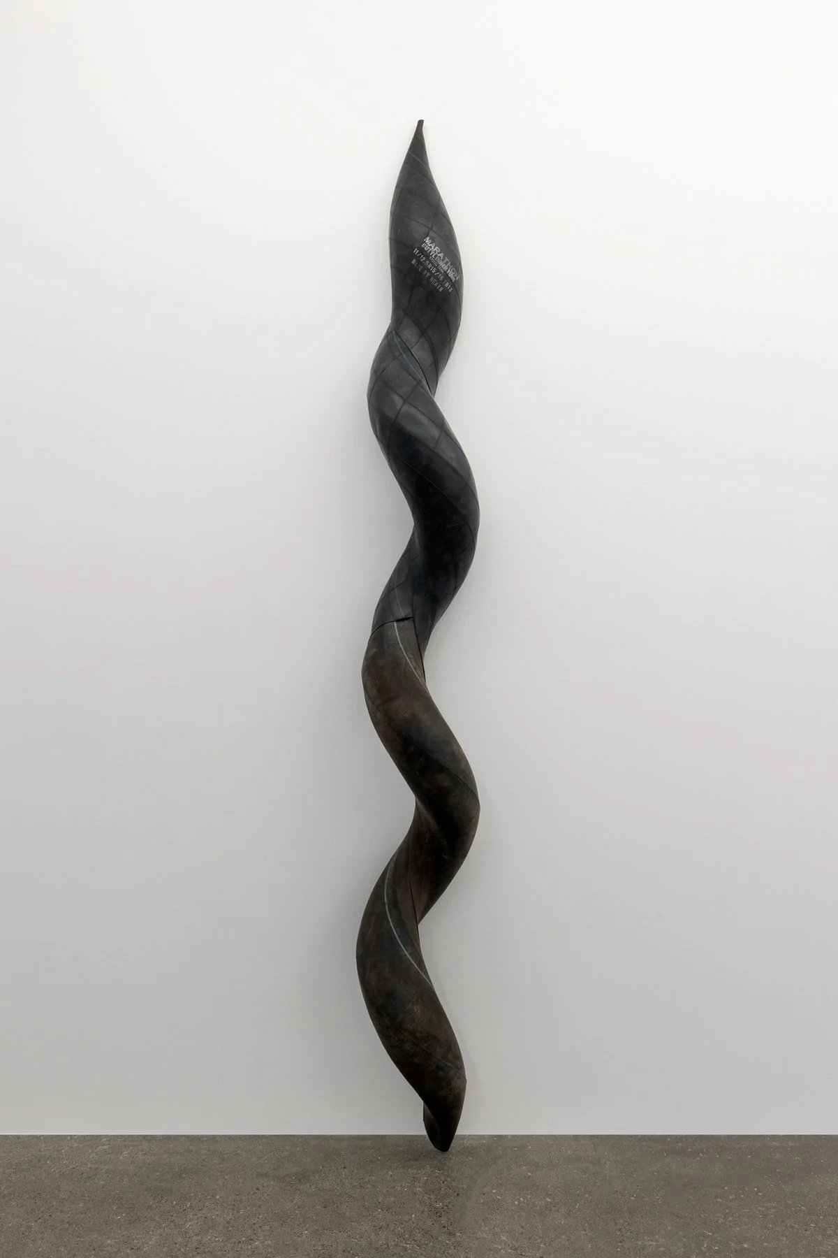 A tall, twisted, black sculpture leaning against a white wall in a gallery.