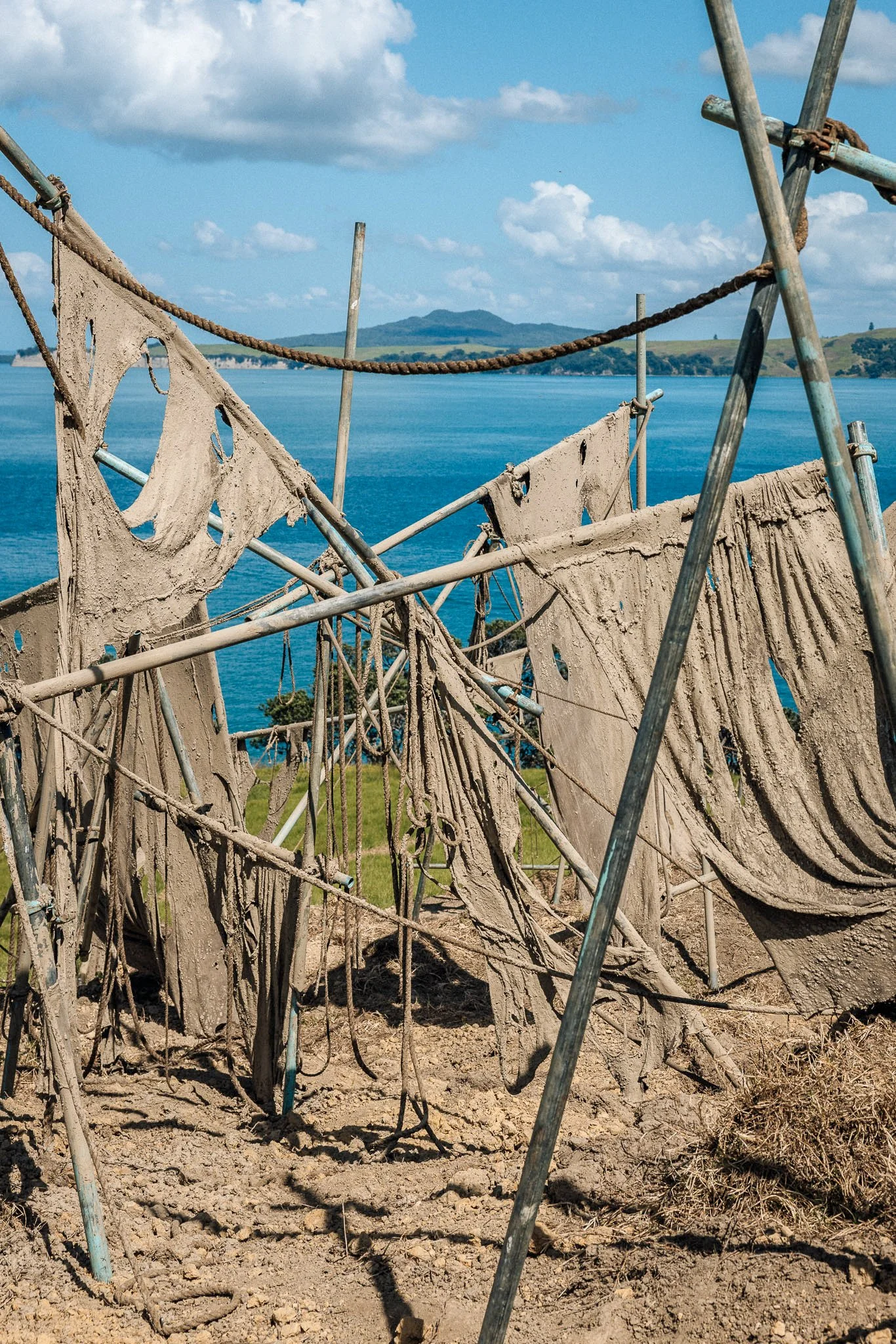 Clothes hanging to dry on a makeshift outdoor drying rack near a body of water, with green landscape and blue sky in the background.
