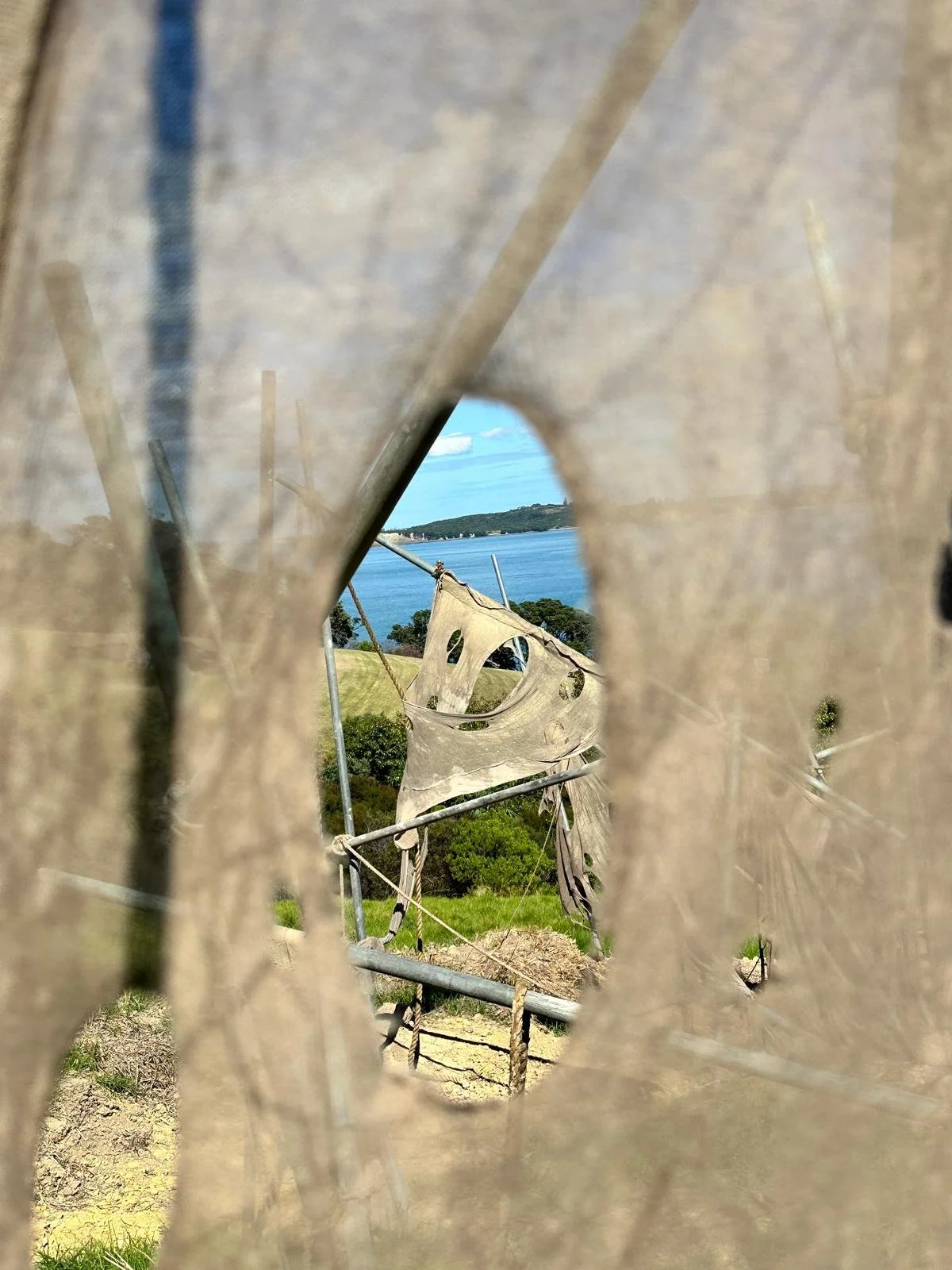 View through a hole in a weathered, torn fabric or tarp to a scenic landscape featuring trees, a body of water, and a distant shoreline under a blue sky.