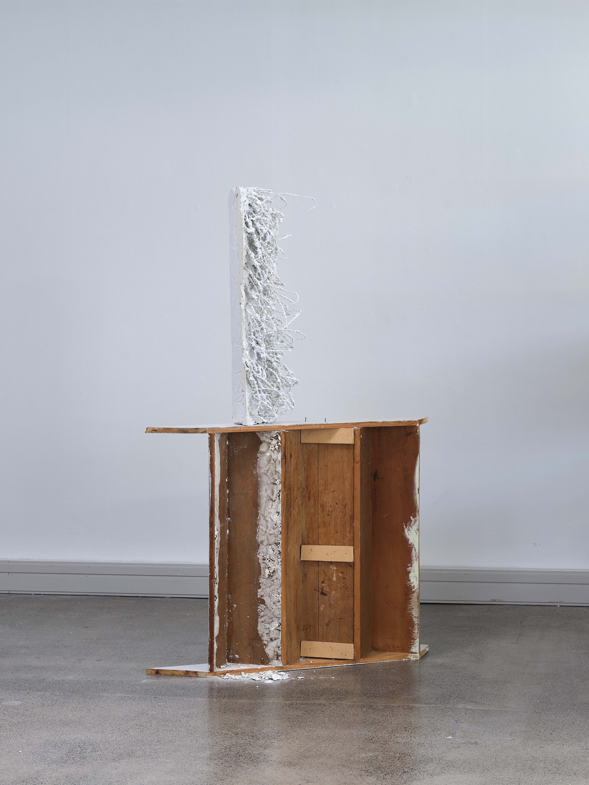 An art installation composed of a partially assembled wooden structure with some white material dripping down from a vertical panel on the left, set against a plain white wall and a gray floor.