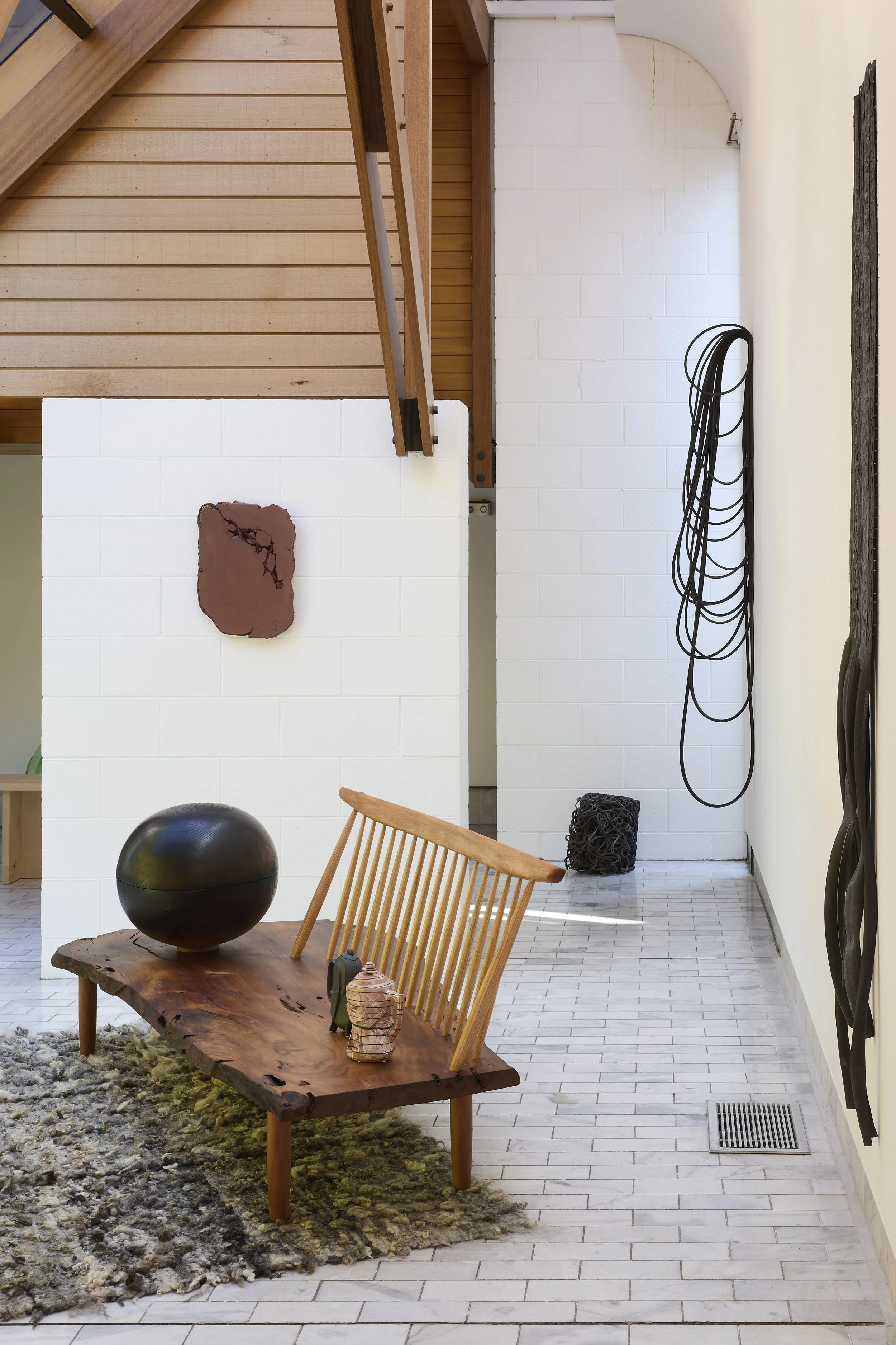 Interior space with wooden staircase, white brick walls, a wooden bench with decorative objects, a black sculpture, and hanging black cords.