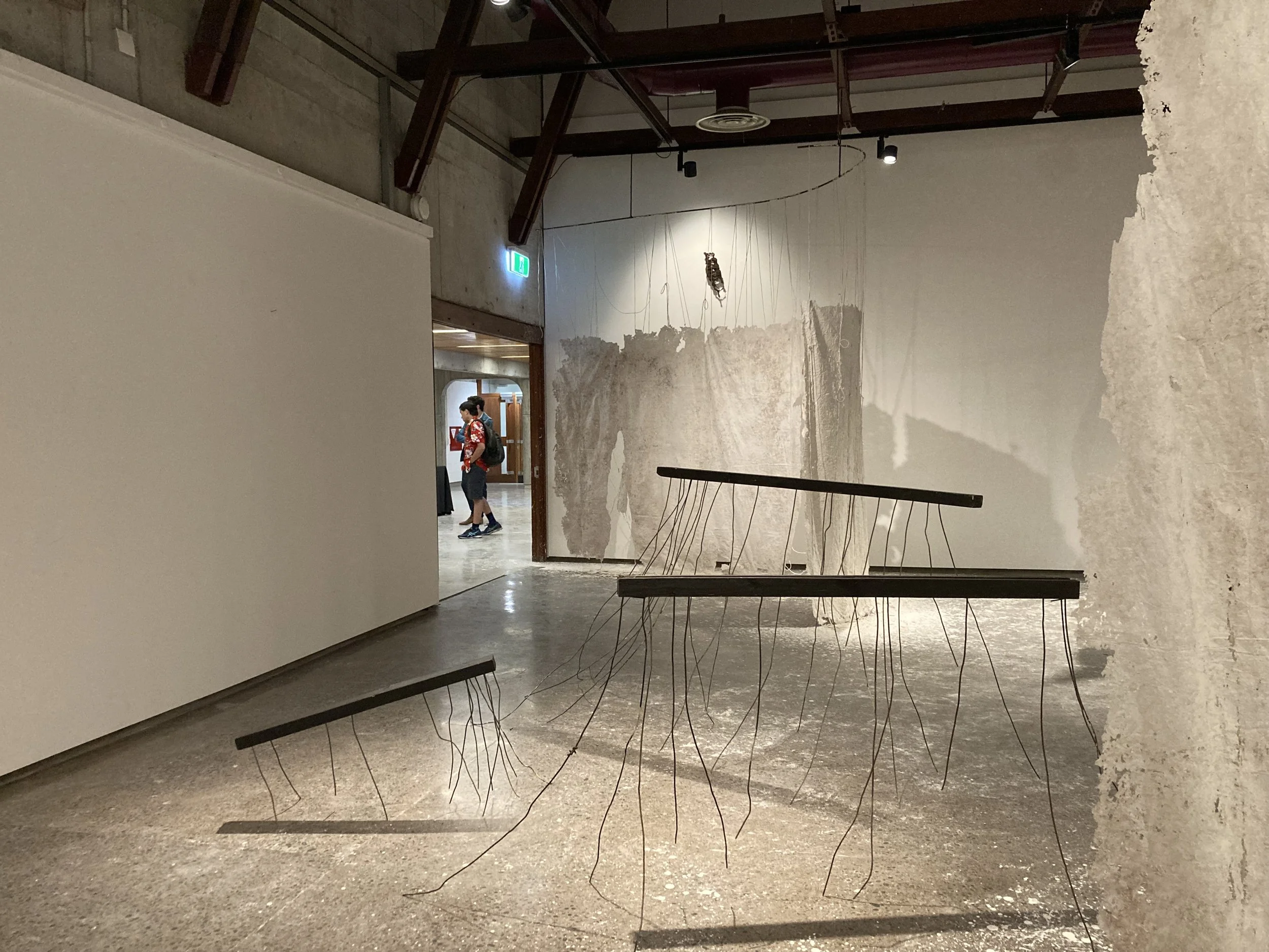 Art installation with three suspended black horizontal bars, each with hanging wires, set against a textured wall with patches of exposed concrete and painter's tape, in a gallery space with concrete floors and an open doorway.