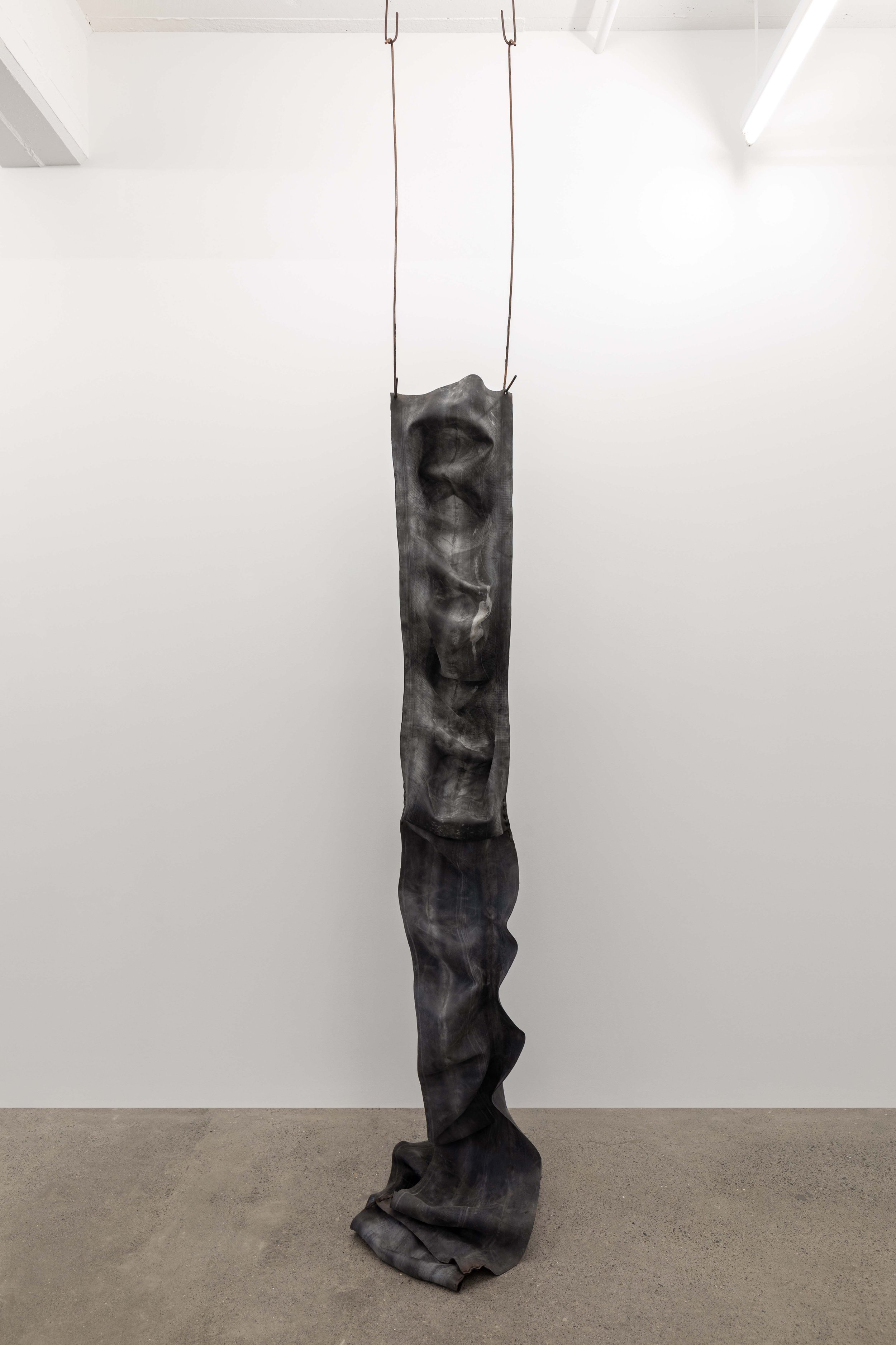 An art installation featuring a long, vertical, dark fabric with a distorted face printed on it, hanging from the ceiling by two hooks.