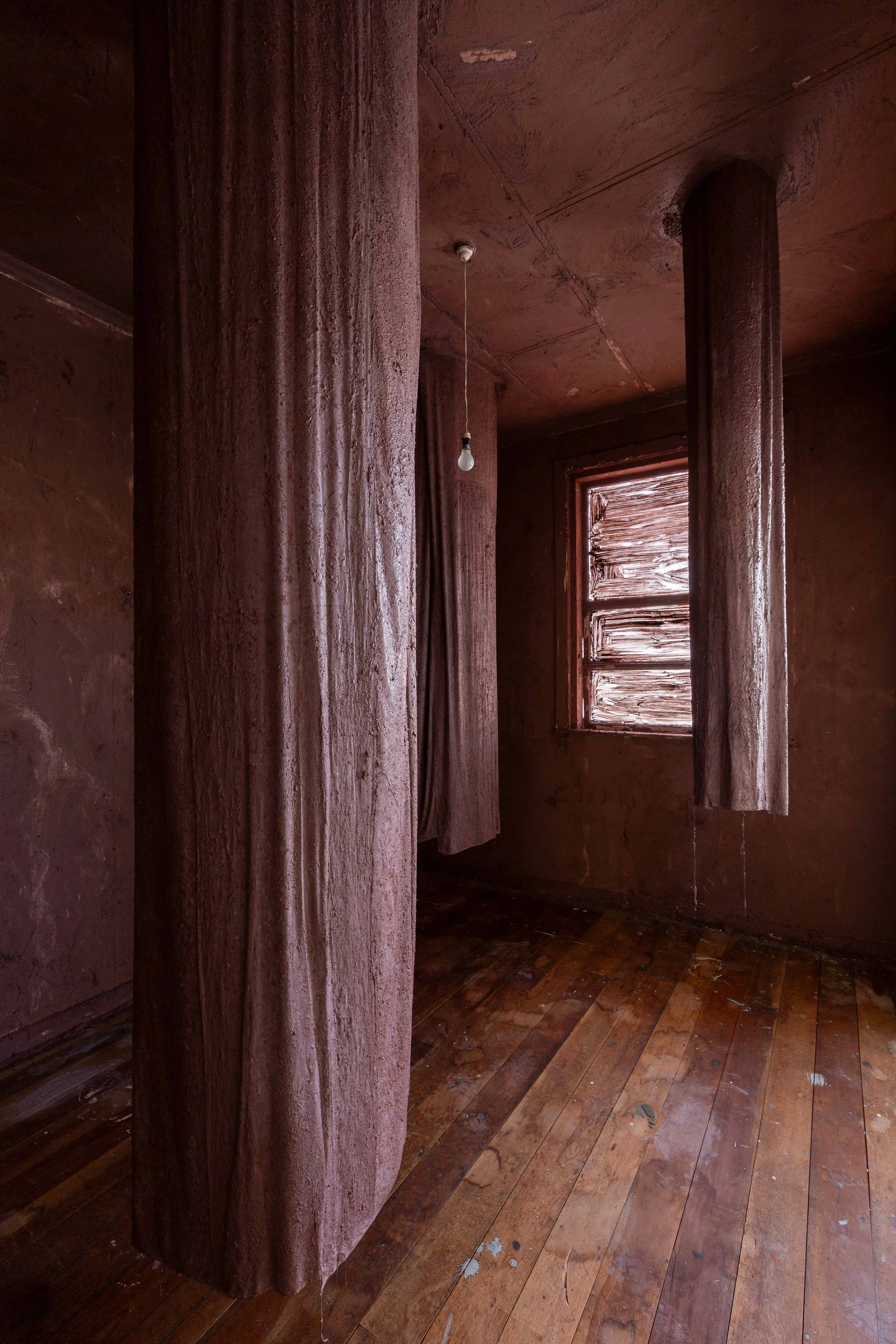 An empty room with reddish-brown walls and wooden floor, with two cylindrical reddish-brown support columns, a window with wooden slats and faint curtains, and a hanging light bulb.