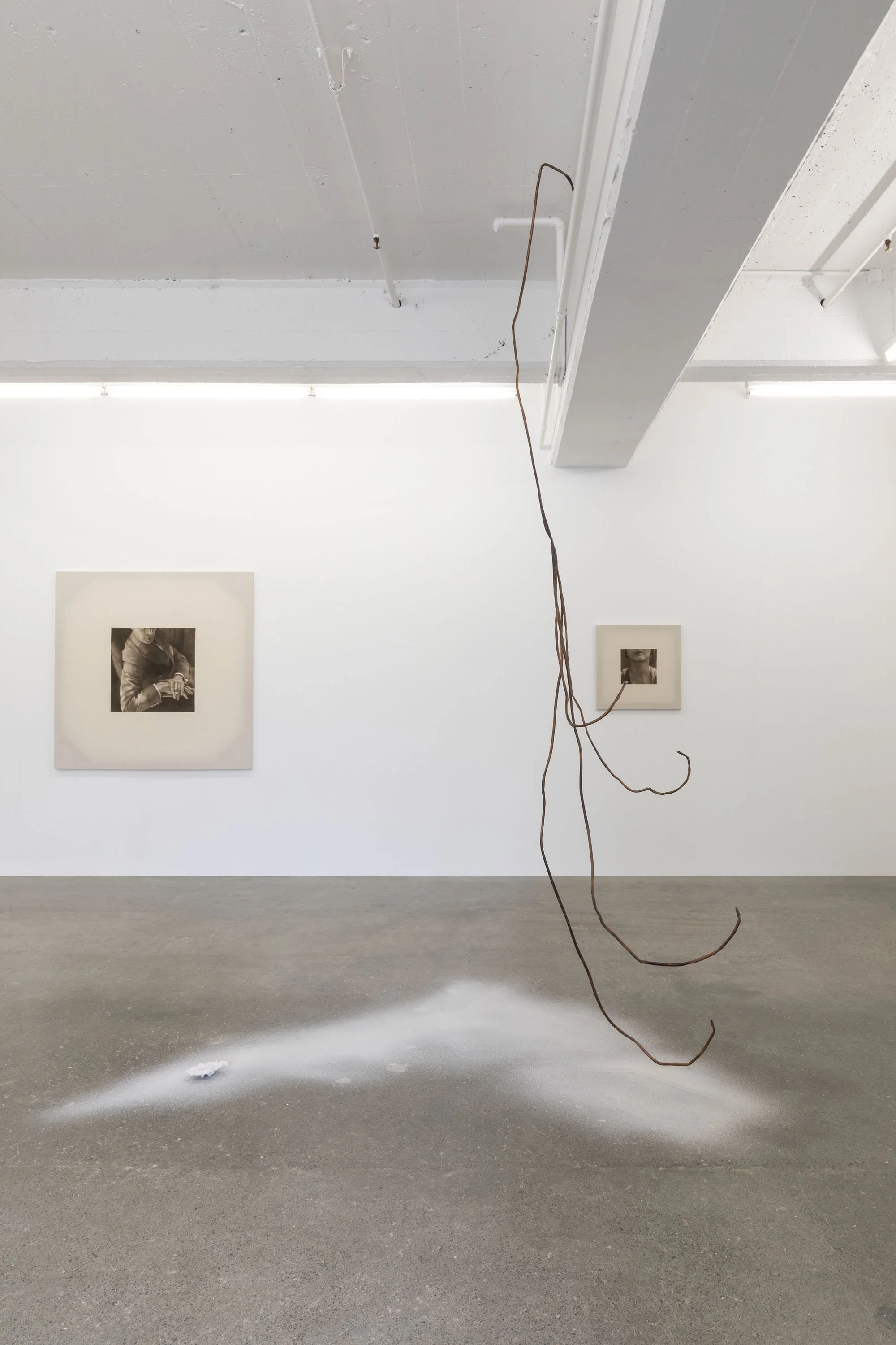 An art gallery with a suspended, rust-colored wire sculpture hanging from the ceiling. The concrete floor has a white powdery substance on it, with a small black object in the powder. Two black and white photographs are displayed on the white gallery wall.