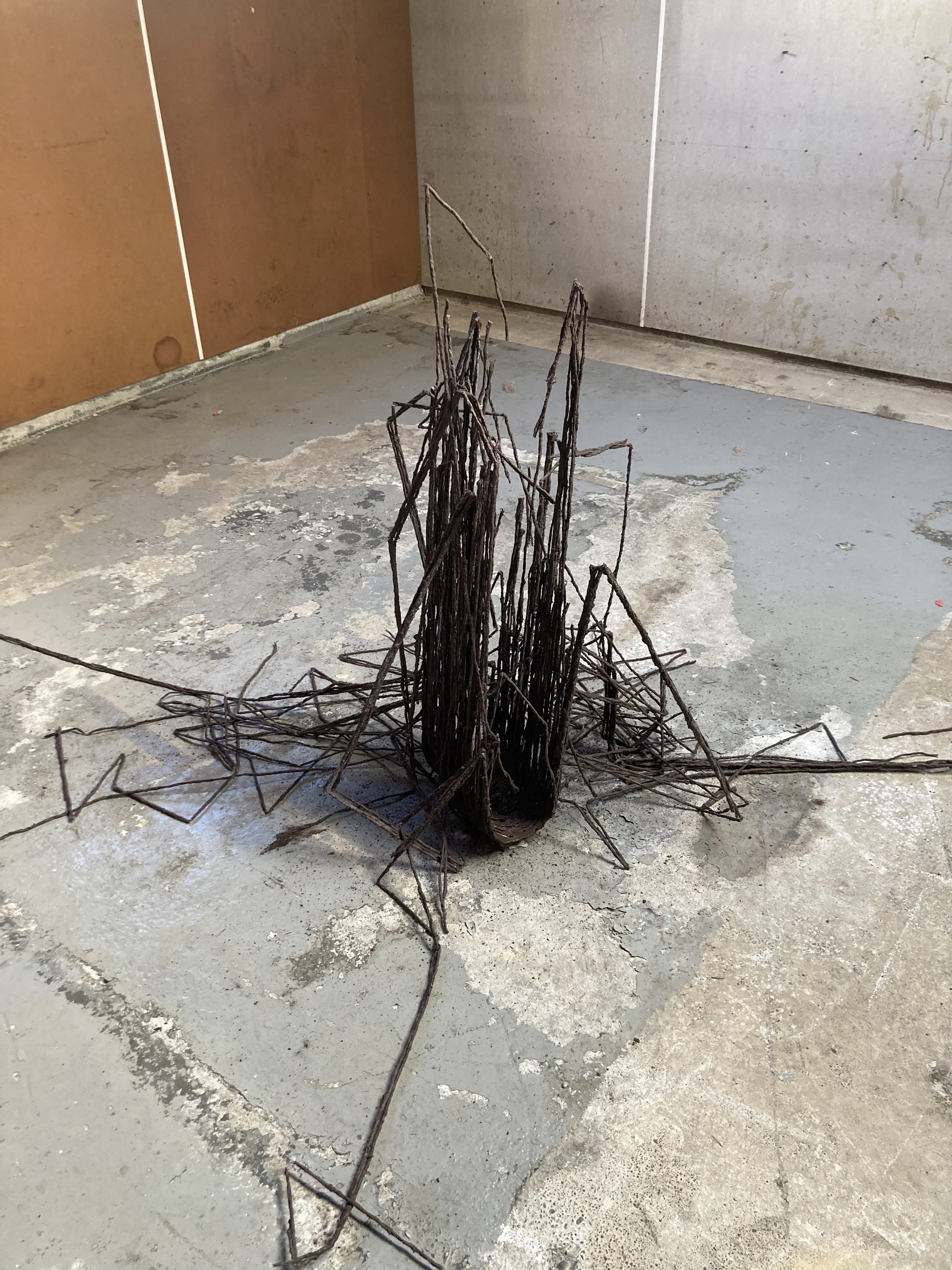 A sculpture made of rusted metal wires resembling a tall, jagged mountain or spire, placed on a concrete floor in an exhibition space.
