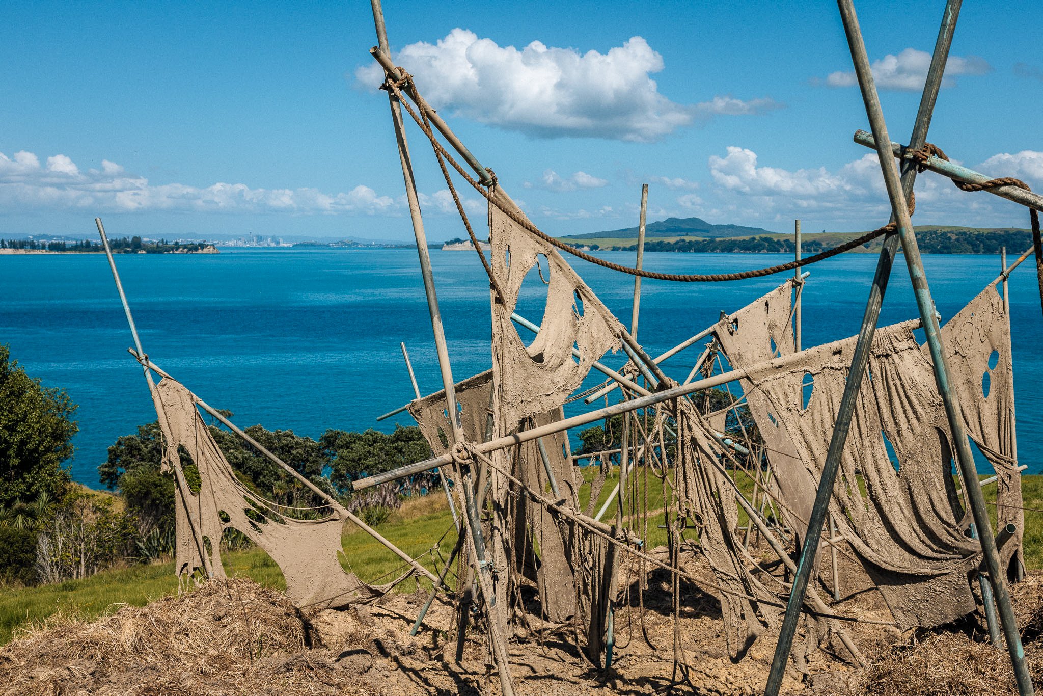 Sculpture resembling a boat made from metal poles and fabric, overlooking a body of water with hills and a partly cloudy sky in the background.