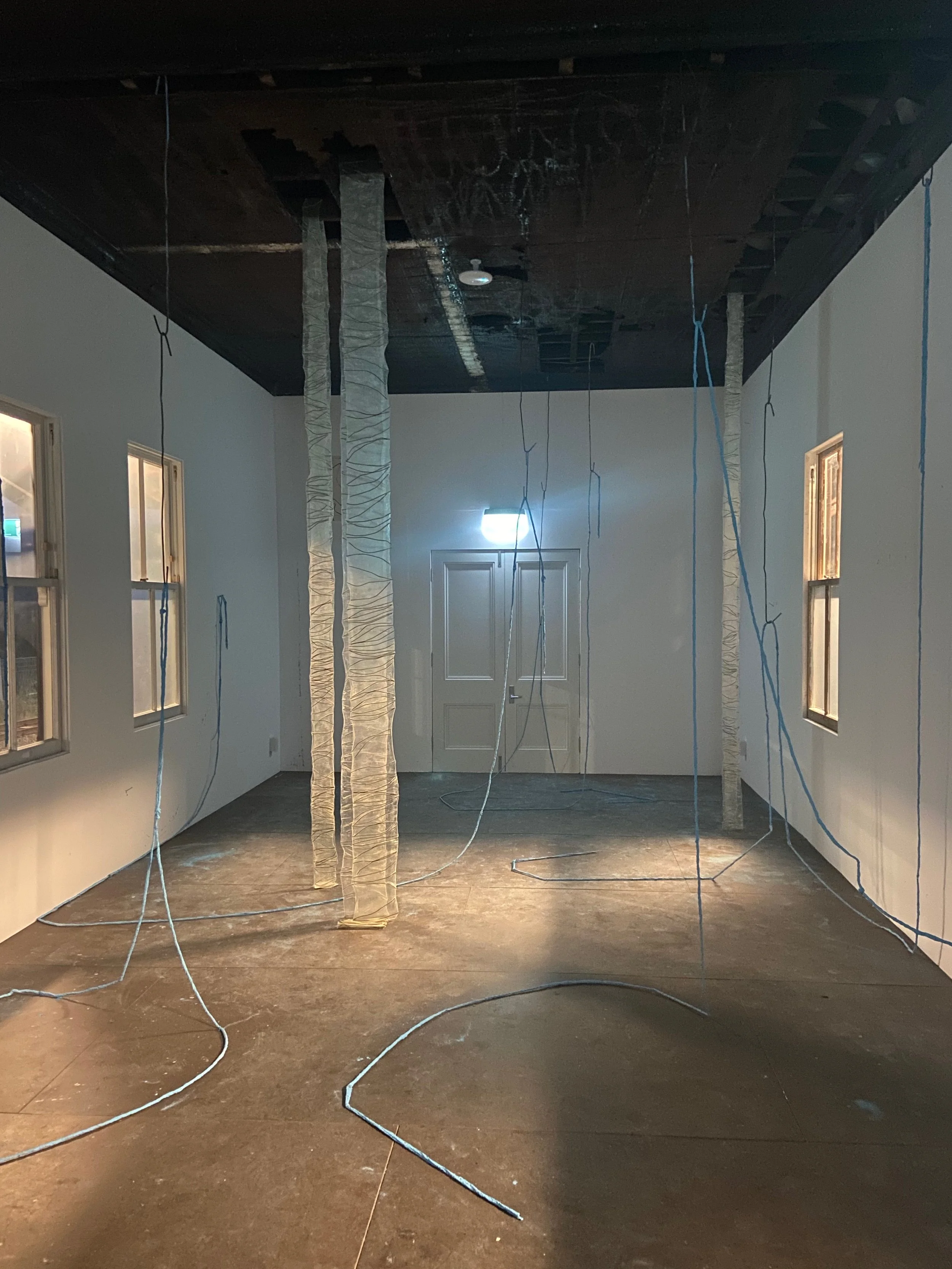Room under construction with wrapped support poles, wires hanging from ceiling, and a door at the back illuminated by a wall light.
