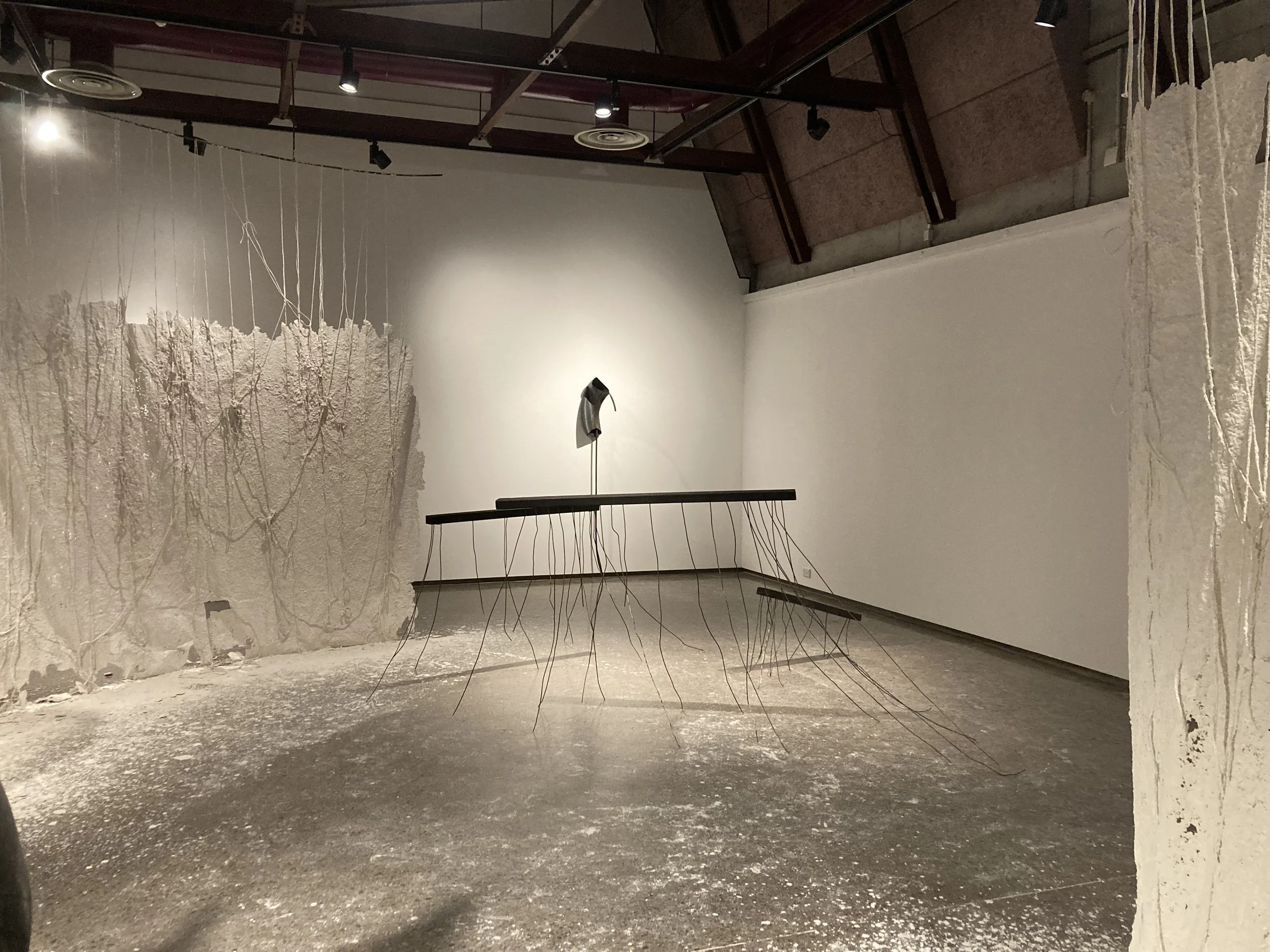 Art installation in a gallery with a textured white wall, thin hanging wires, a suspended object that looks like a mask, and a long horizontal surface supported by wires, with a concrete floor.