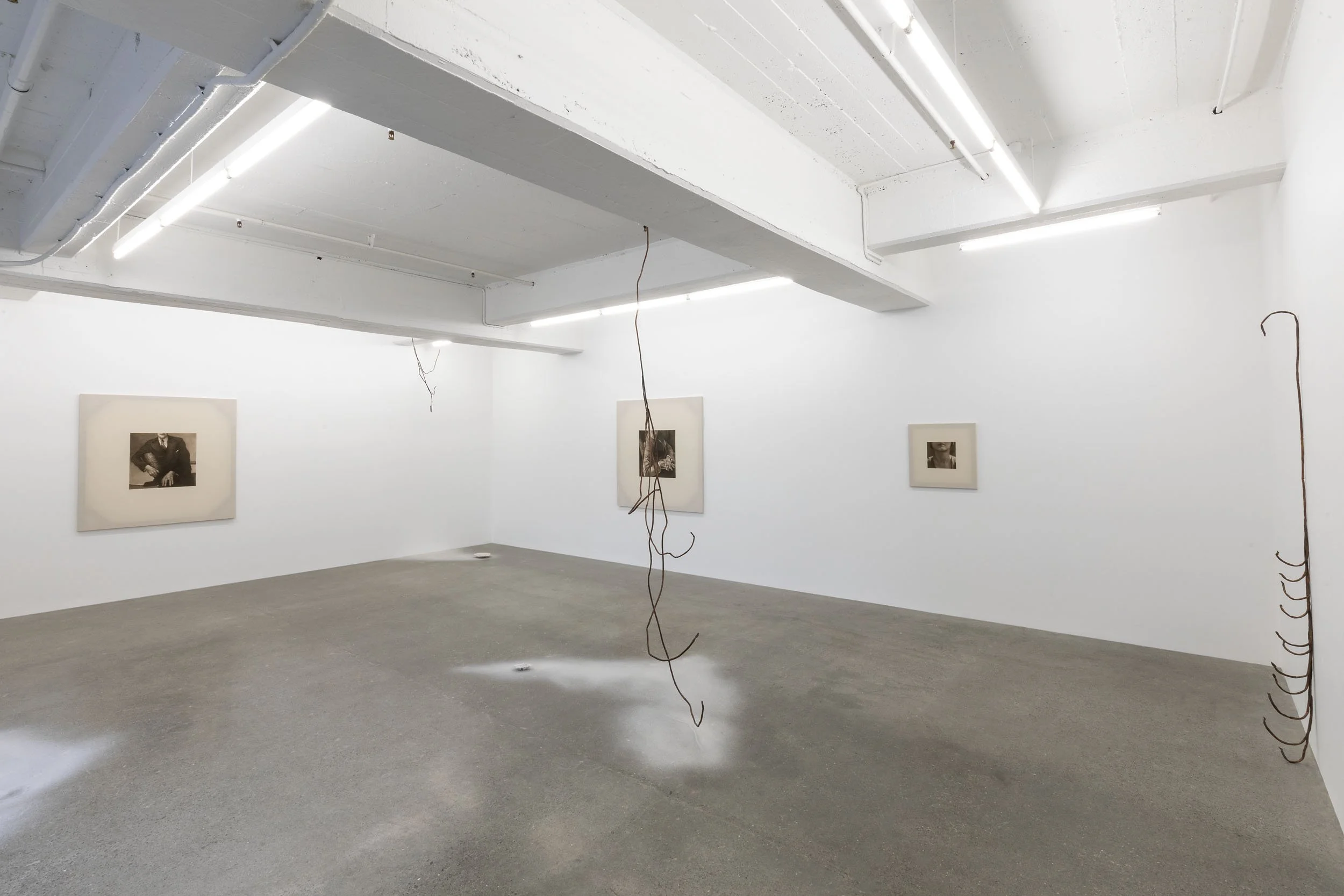 An art gallery with white walls, concrete floor, and framed black-and-white photographs on display. There are metal sculptures hanging from the ceiling and touching the floor, with some ink-like splatters on the ground.