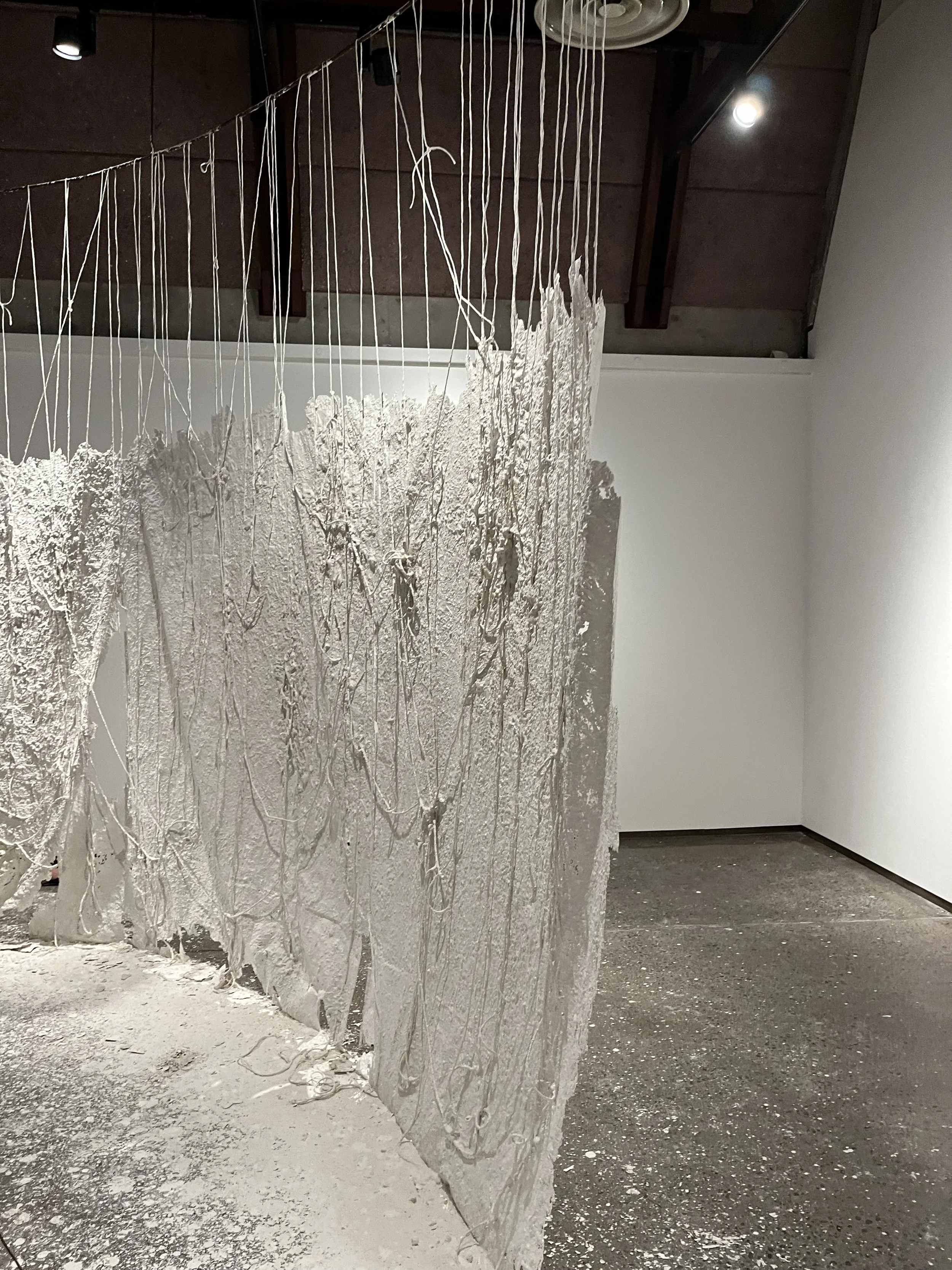 An art installation featuring a textured, uneven, white surface with hanging threads or cords, set in a gallery space with plain white and wooden walls and concrete floors.