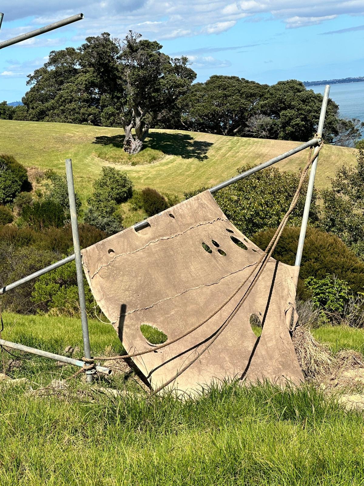 A large, weathered piece of fabric with multiple holes, supported by metal poles and ropes, is set on green grass in a hillside landscape with trees and a view of the ocean in the background.