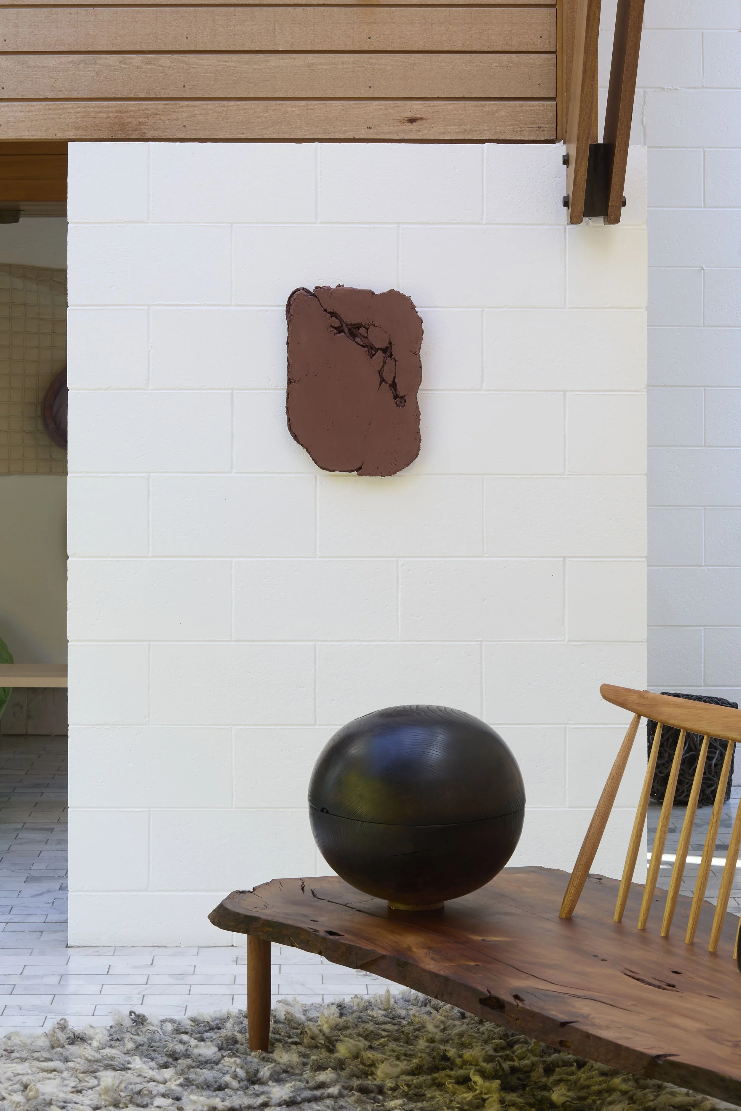 Photograph of a modern interior with a white brick wall, a black spherical sculpture on a wooden table, and a wooden chair with vertical slats. A piece of abstract wall art hangs on the wall above.