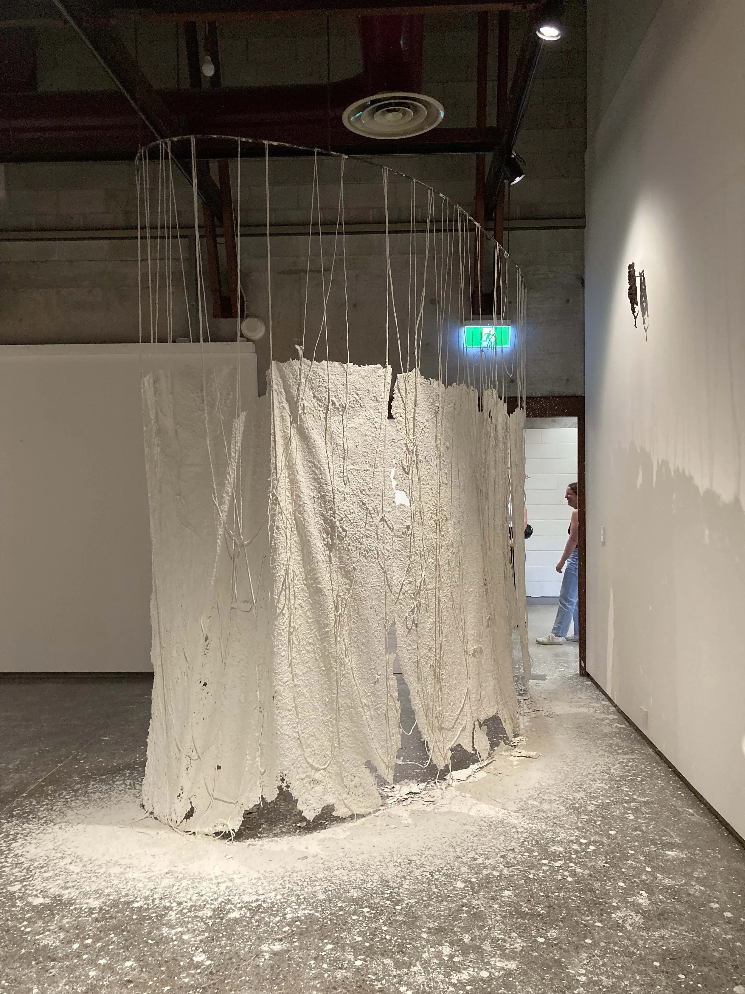 Contemporary art installation featuring a circular structure made of textured white material, suspended by numerous strings from the ceiling, with powder or dust scattered on the floor beneath it.