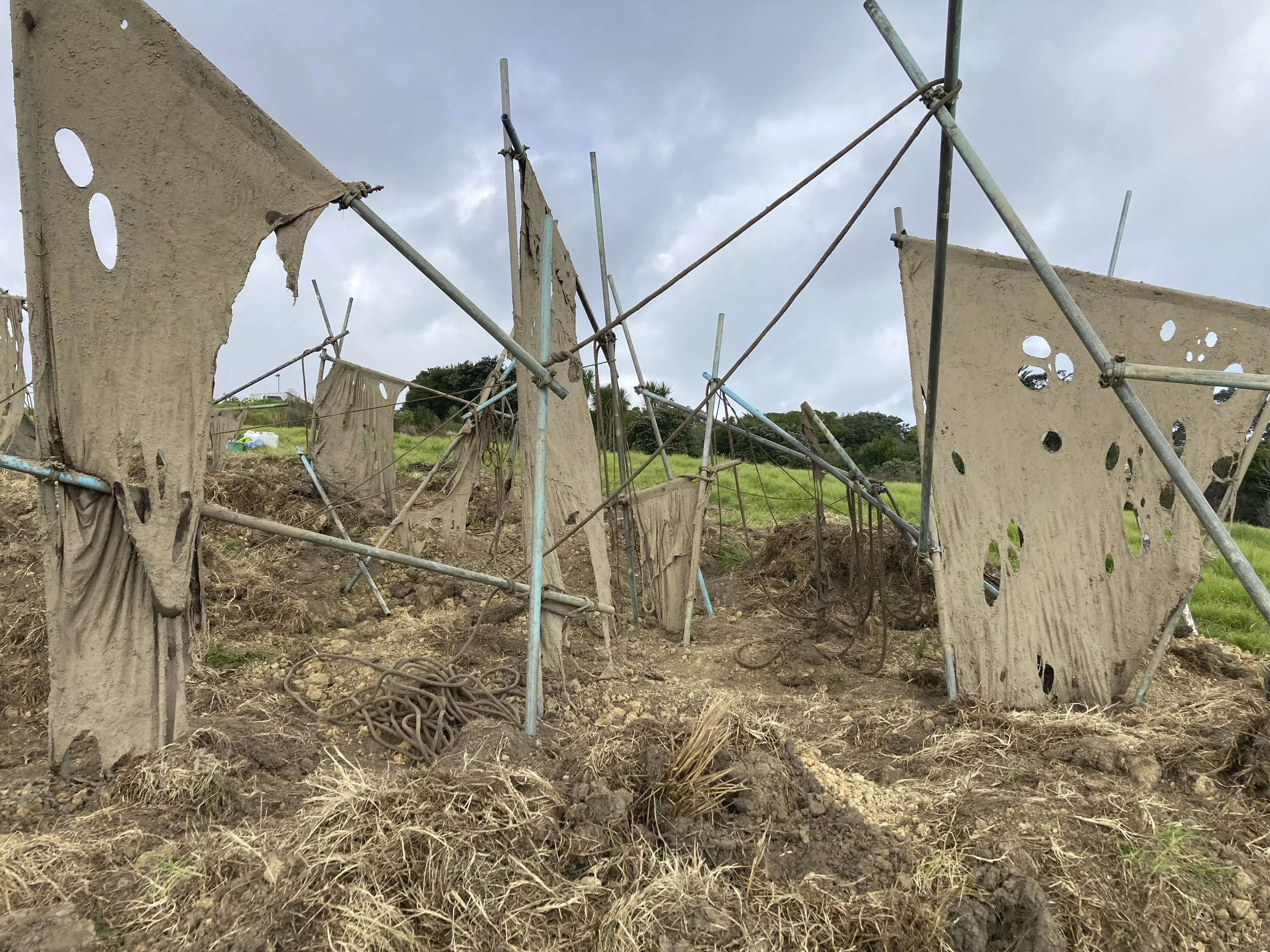 clothes drying rack with tattered, holey fabrics hanging in an open field with green grass and cloudy sky