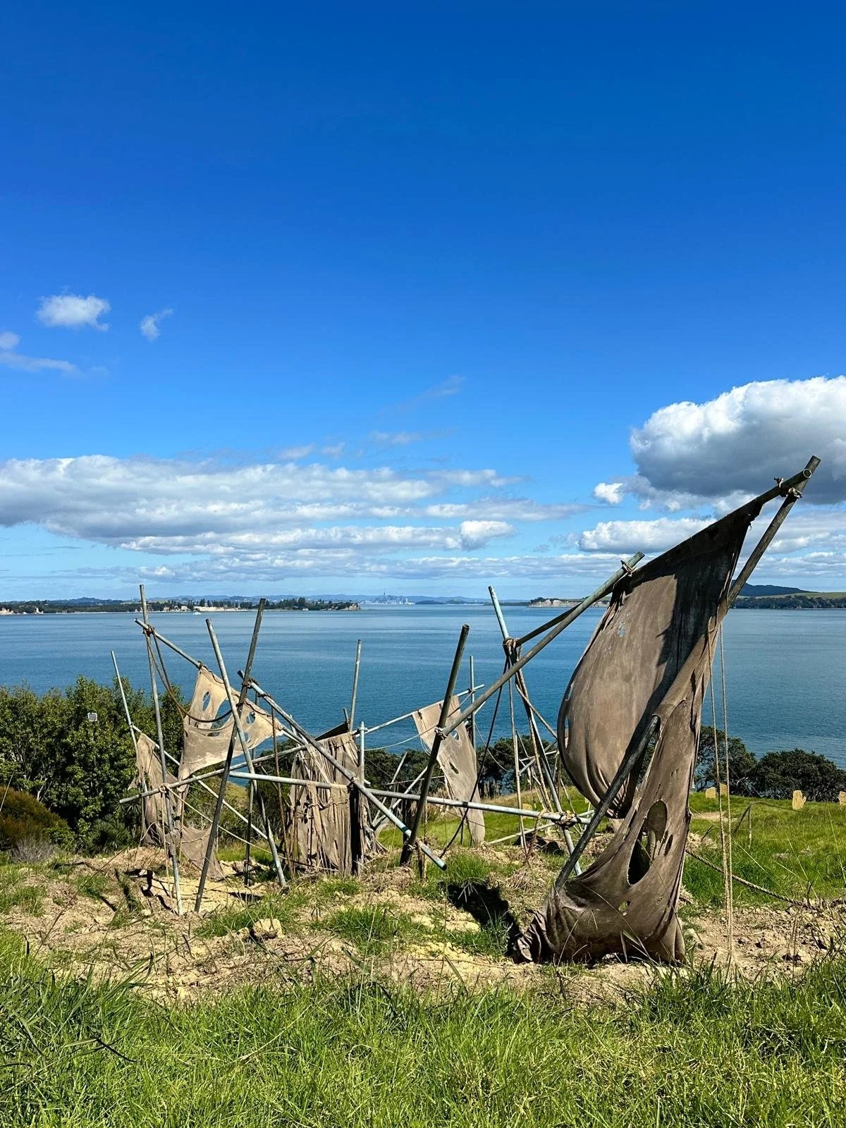 Scenic view of an art installation with weathered fabric and metal poles on a grassy hill overlooking a body of water with distant land and a partly cloudy blue sky.