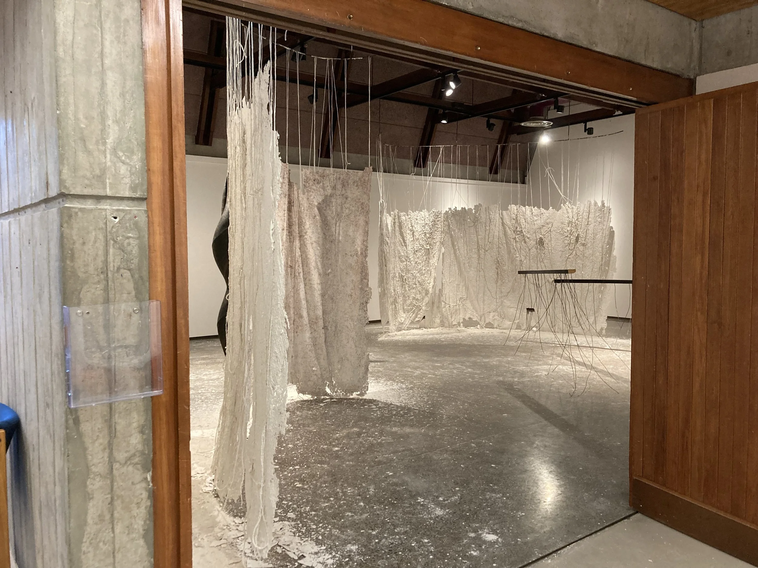 An art installation in a gallery featuring large, textured, white sculptural pieces with hanging threads and a suspended fabric sheet, with spotlights illuminating the artwork.