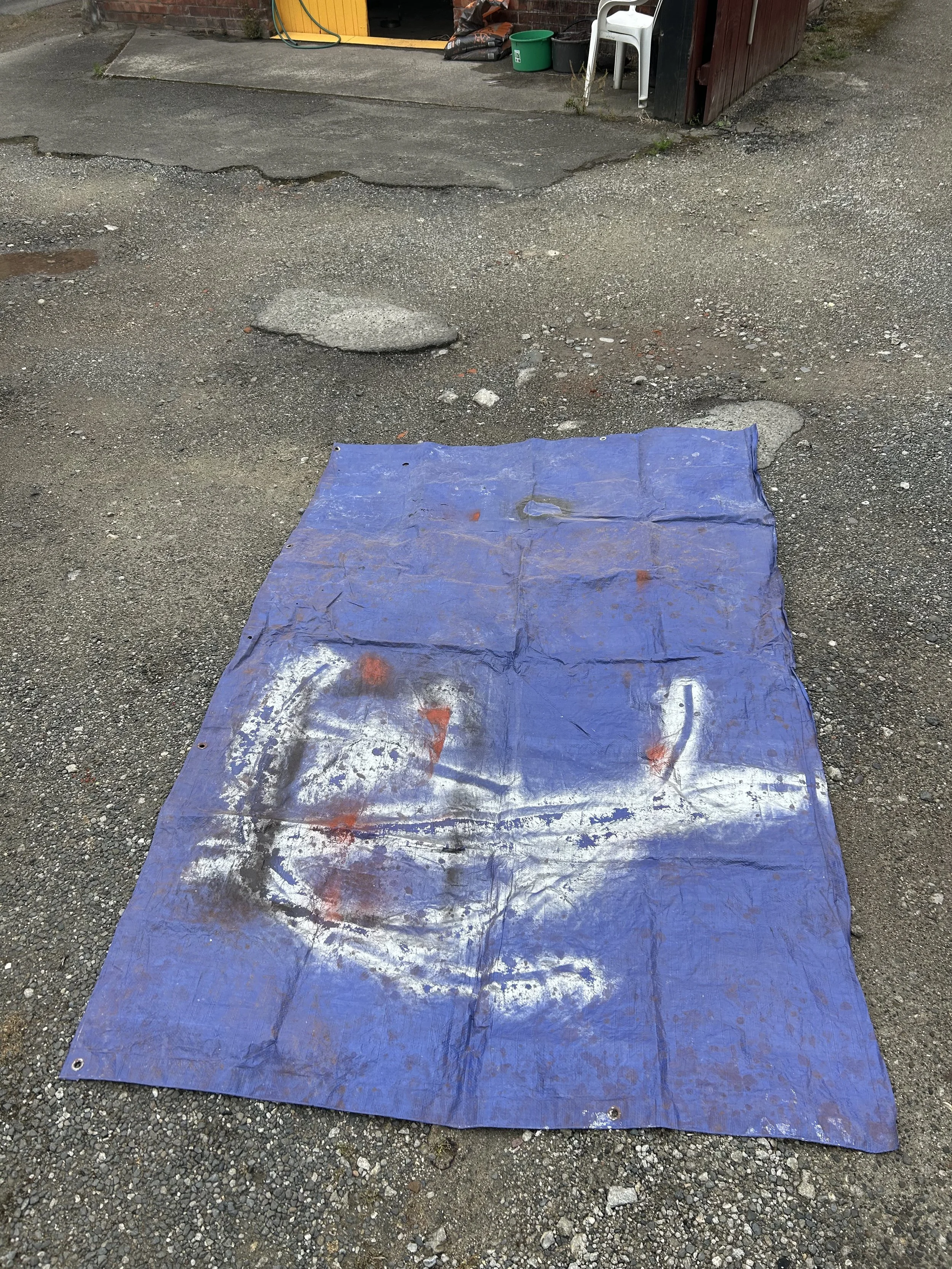 A blue tarp on a gravel ground with spray-painted white and red markings, with a background showing part of a building, a green bucket, a white chair, and some objects near a brick wall.