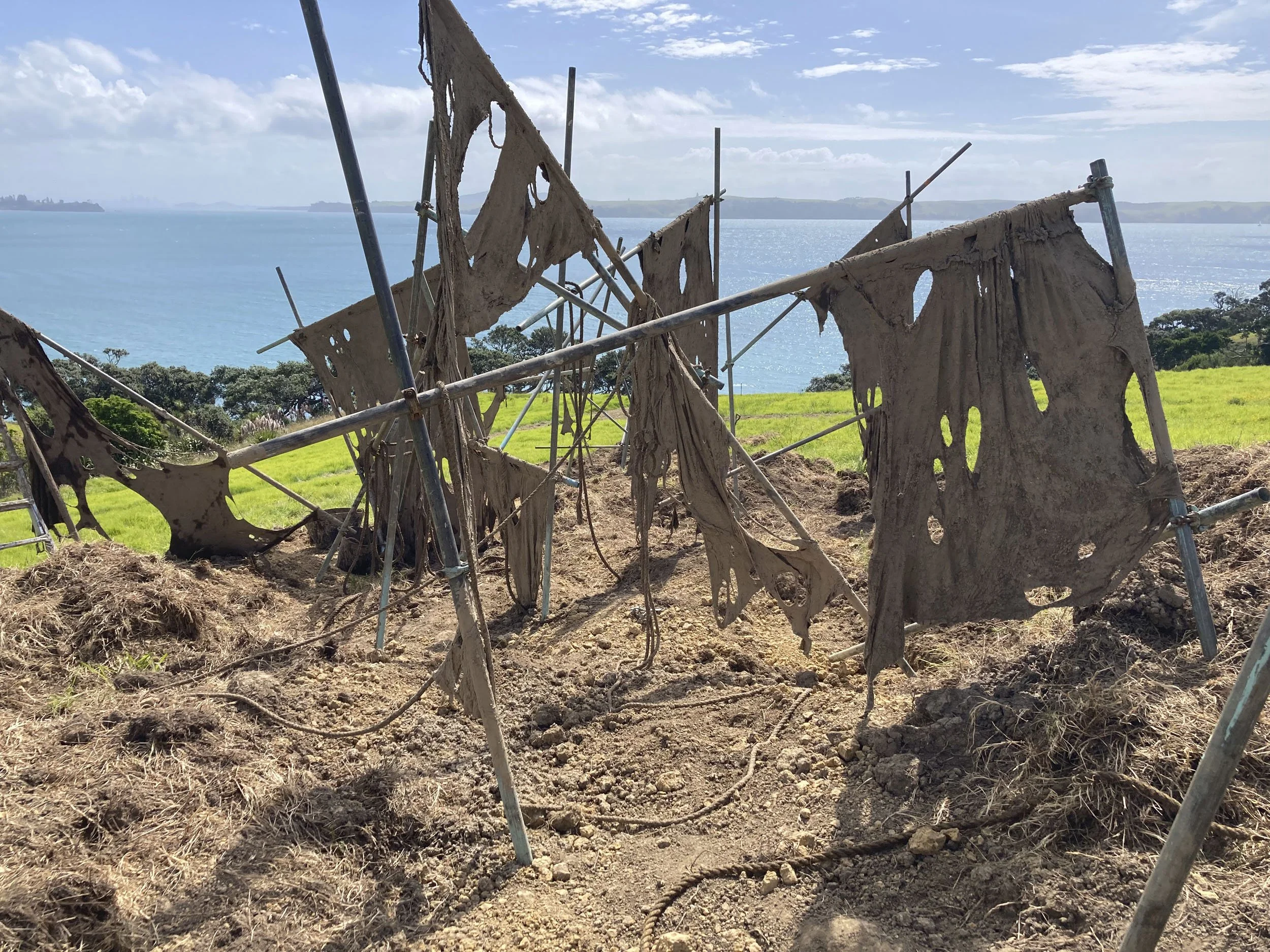 Tattered and torn fabric panels hanging on a metal structure overlooking green fields and water.