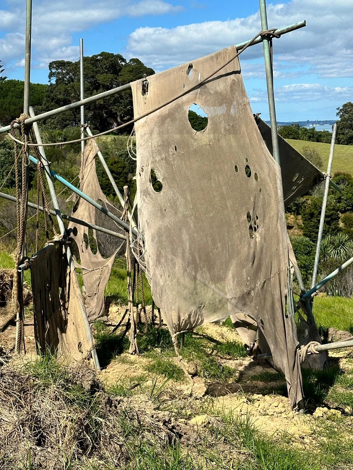 Tattered, weathered cloth hung on a metal structure, with a landscape of trees, grassy hills, and blue sky with scattered clouds in the background.