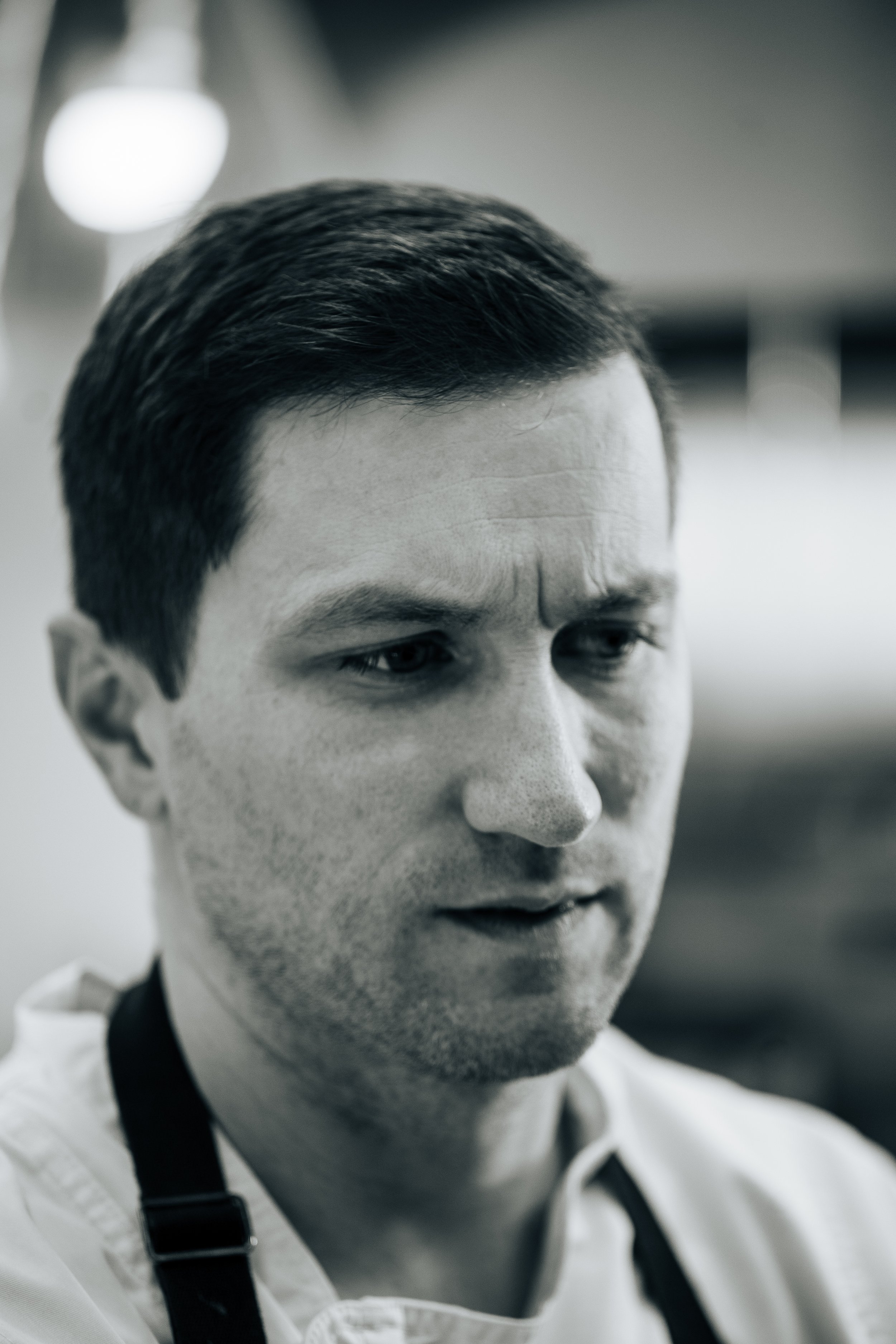 Black and white close-up of a man with dark hair, wearing a white shirt and a black apron, with a focused expression.