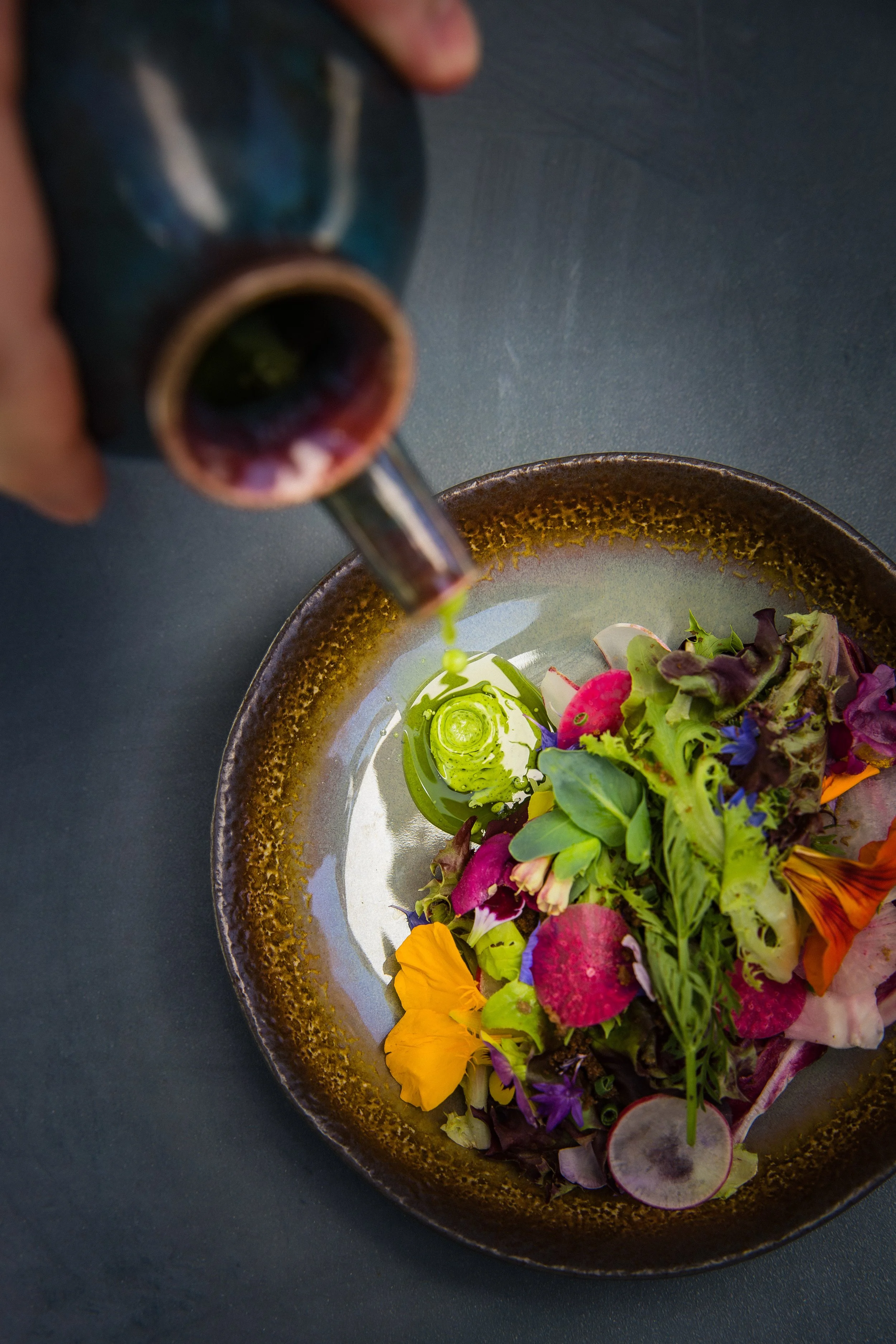 A colorful salad with edible flowers, radishes, and greens on a black plate, being drizzled with dressing from a soy sauce bottle.