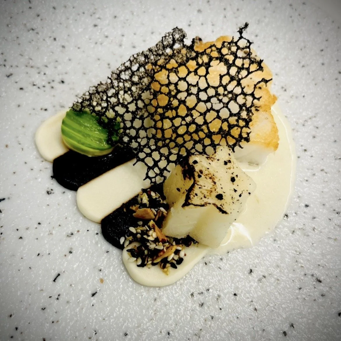 Pan-seared Sea bass rests atop a duet of black and white celery root pur&eacute;es, accompanied by confit celery root and a sesame granola for texture. A delicate black garlic tuile adds depth, while citrus-marinated avocado brings brightness. The di