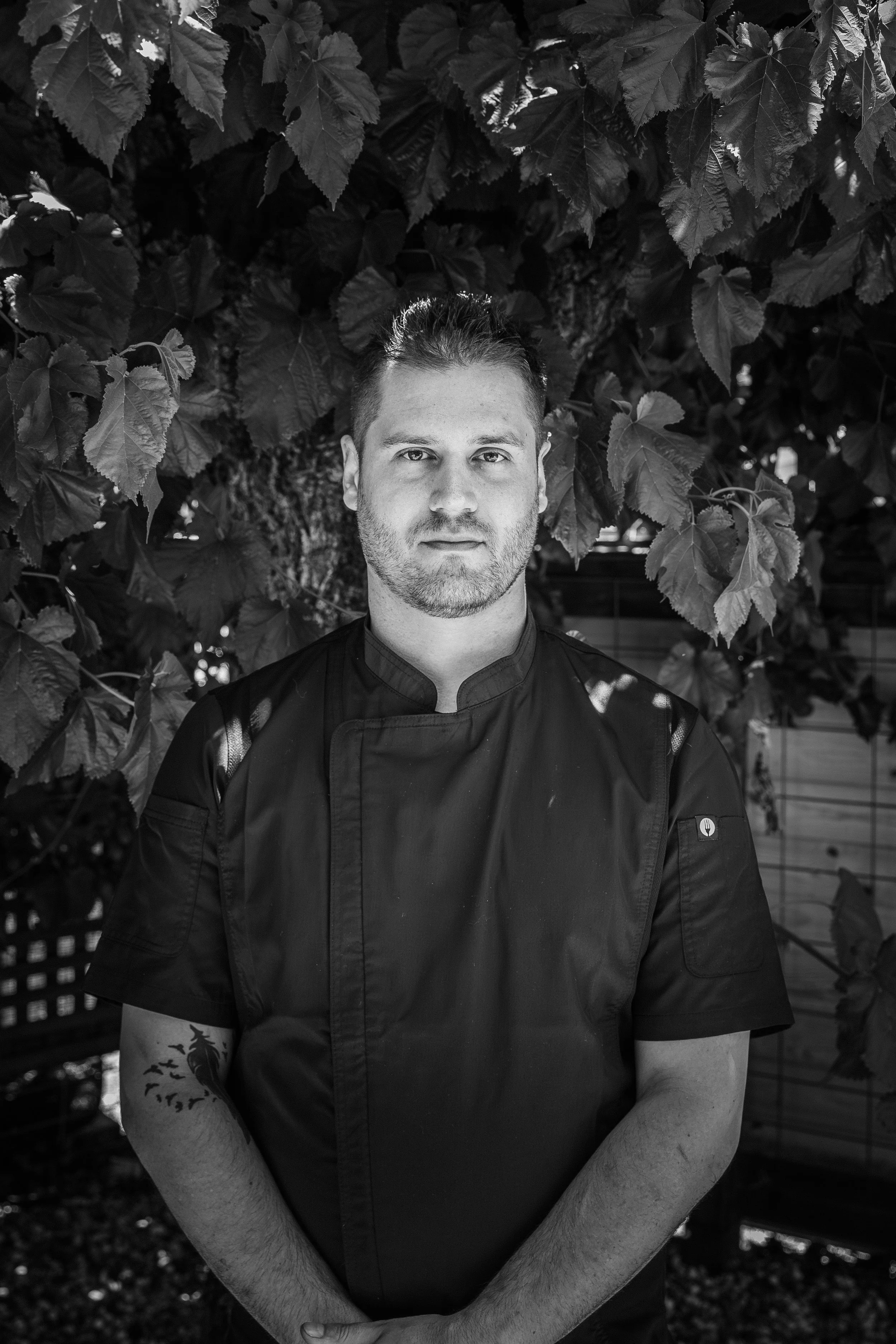 A man in a chef's uniform stands outdoors in front of large leafy plants.