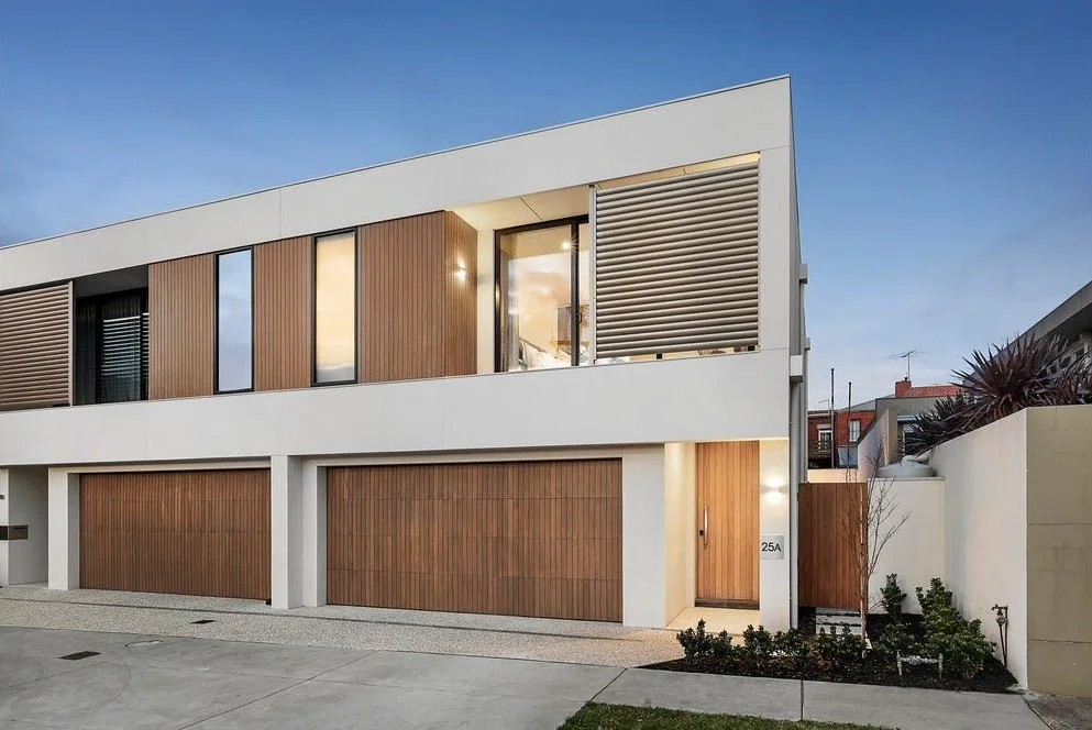 Modern two-story house with white and wood exterior, large windows, garage doors, and a front entrance, during dusk.