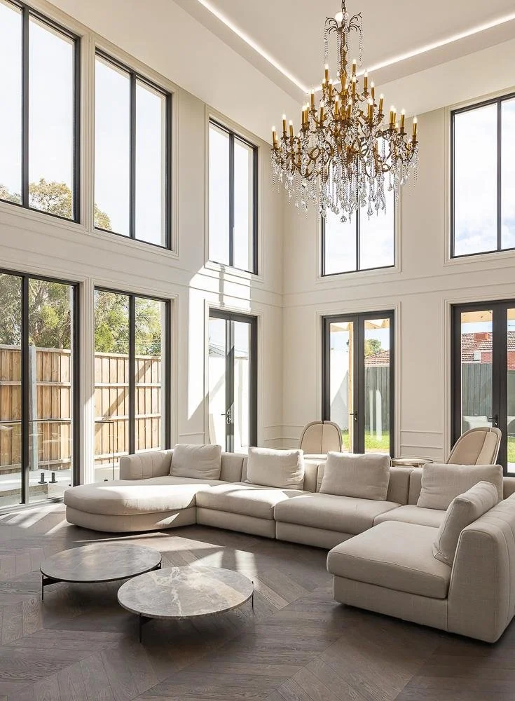 Bright living room with large windows, white sectional sofa, marble-topped coffee tables, and a chandelier hanging from a high ceiling.