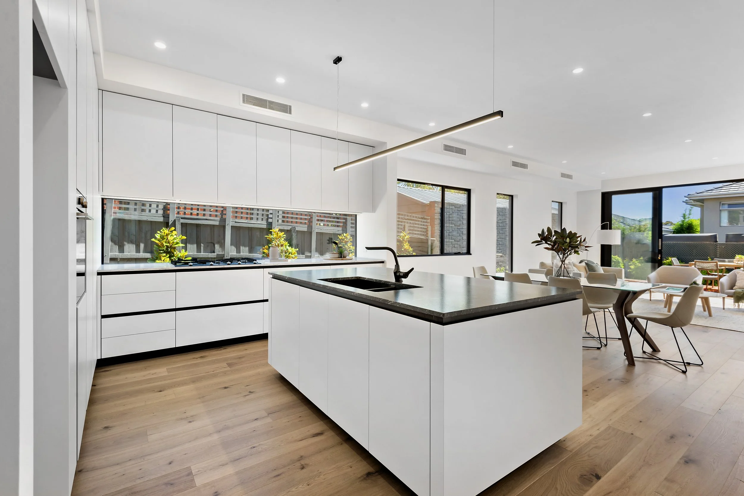 Modern open-concept kitchen with white cabinets, a black countertop island, and a dining area with white chairs and a glass table, featuring large windows and sliding glass door leading to an outdoor patio.