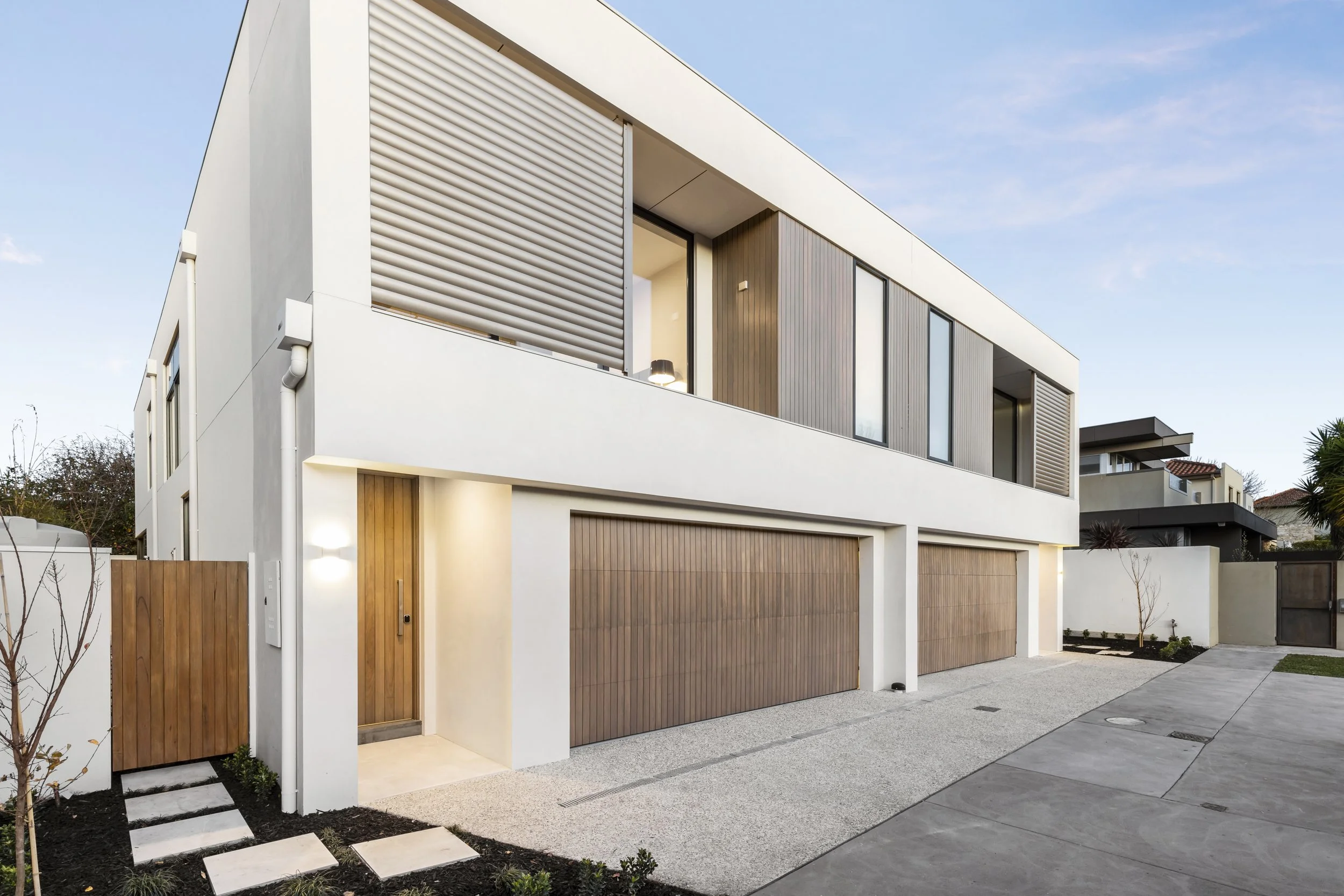 Modern residential building with white walls, wooden garage doors, and a minimalist design, featuring large windows and a small landscaped area in front.