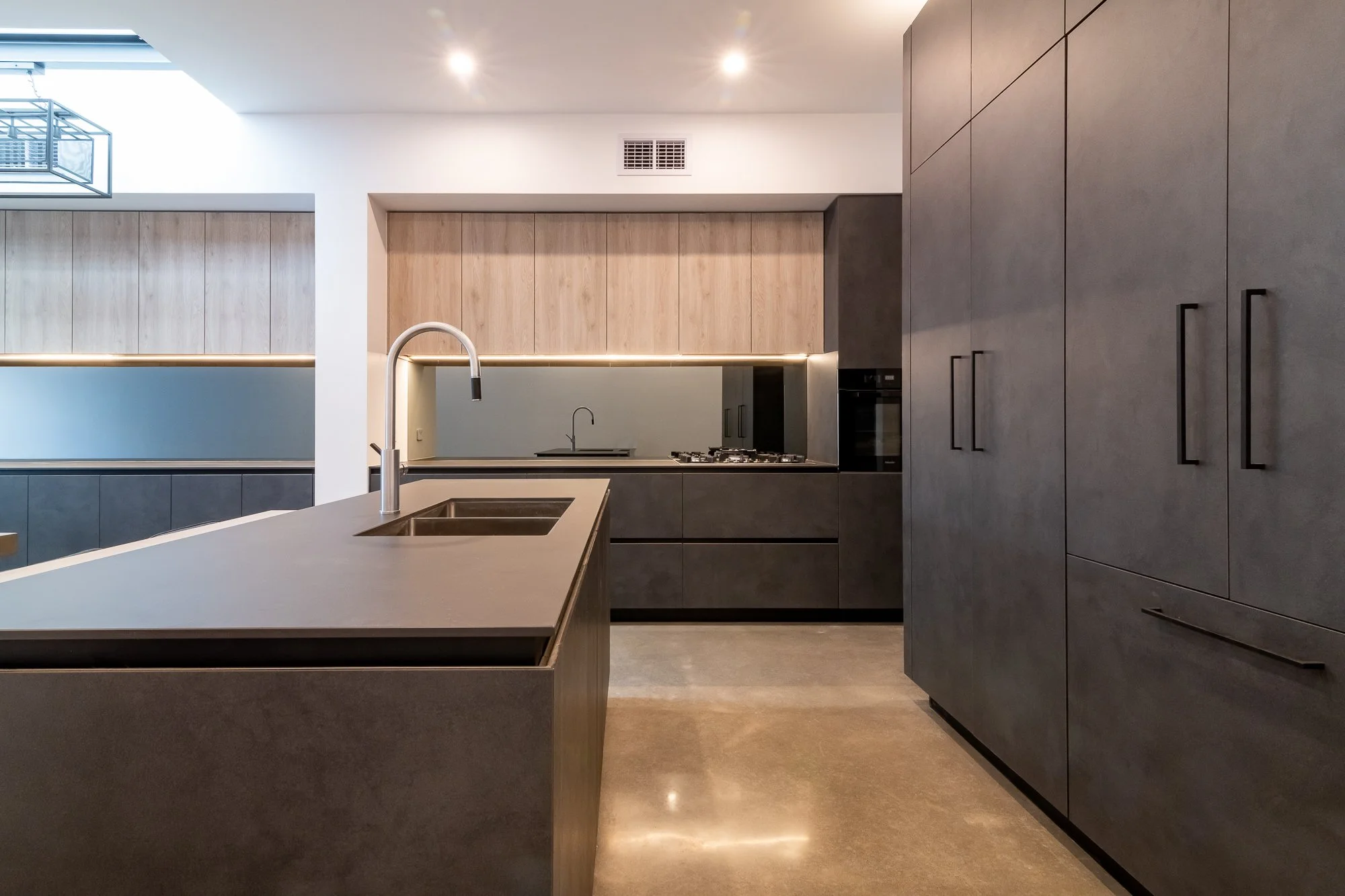 Modern kitchen with dark cabinetry, light wood upper cabinets, a large island with a sink and a modern faucet, stainless steel stove, and built-in oven, with recessed lighting and a reflective floor.