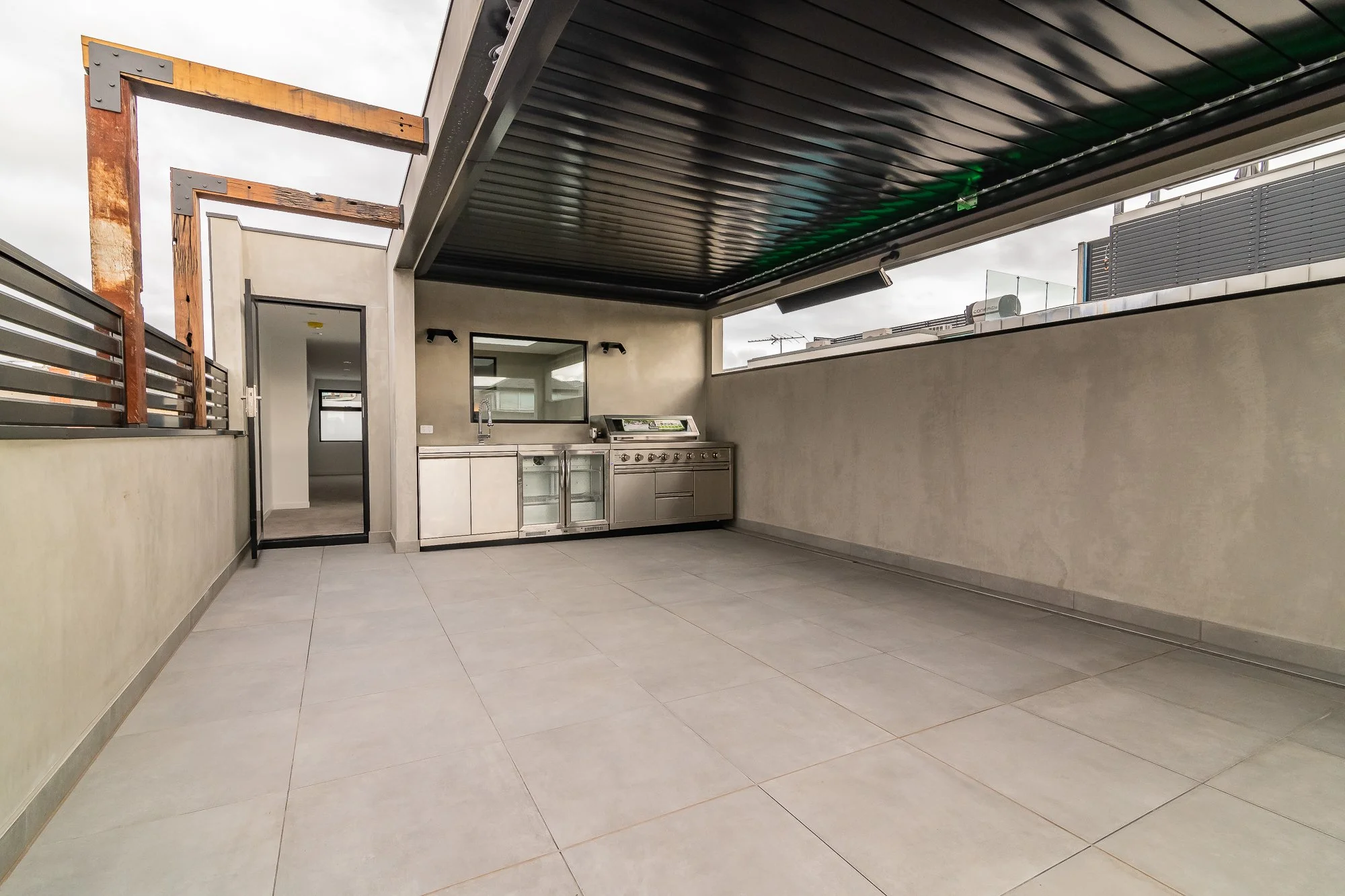 Outdoor rooftop patio with built-in grill, stainless steel kitchen sink, small refrigerator, and open space, enclosed by a wall with a door leading inside and a small window.