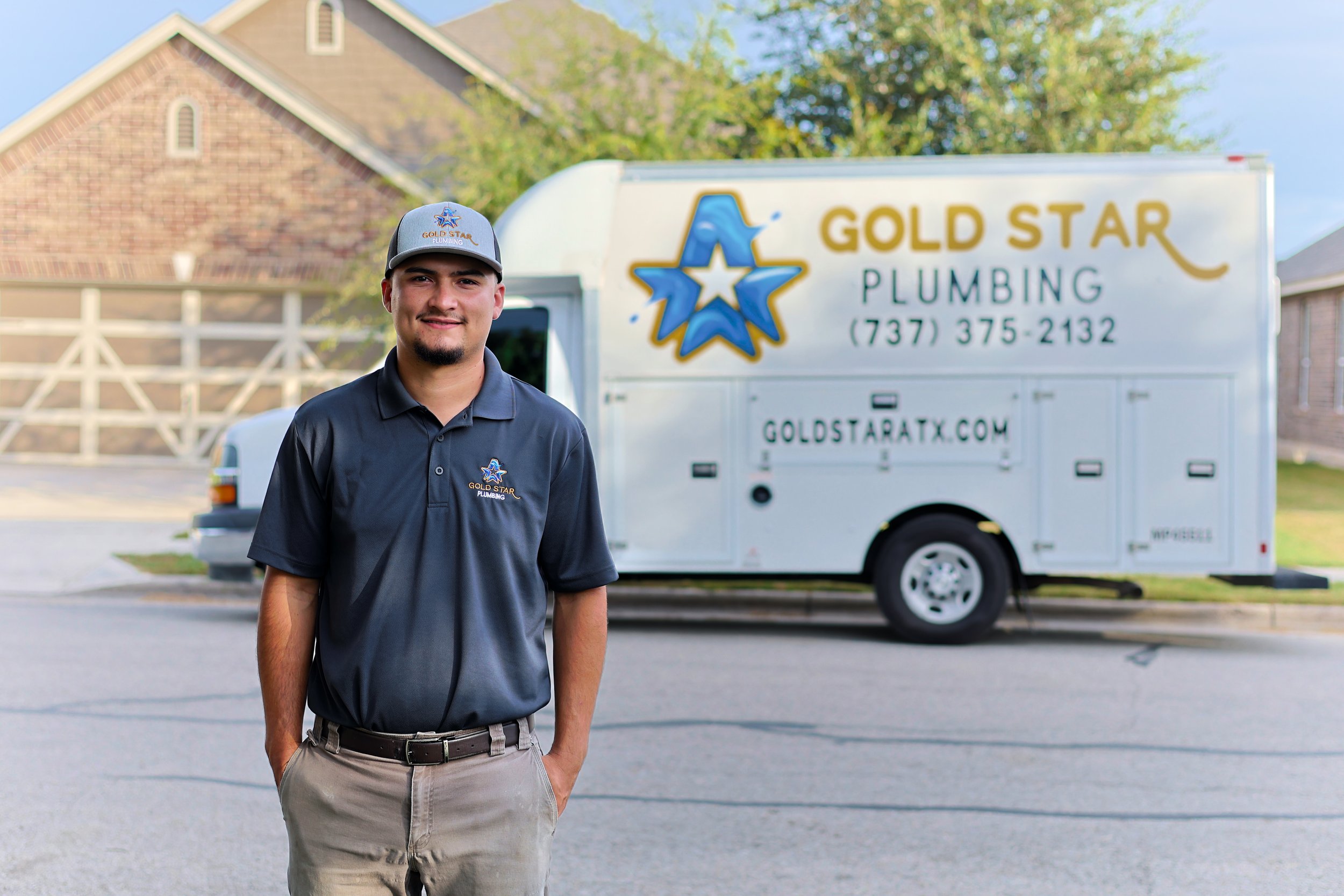 A smiling man in a dark gray polo and baseball cap standing in front of a white plumbing service truck with the logo 'Gold Star Plumbing' and contact information, parked on the street near residential houses.