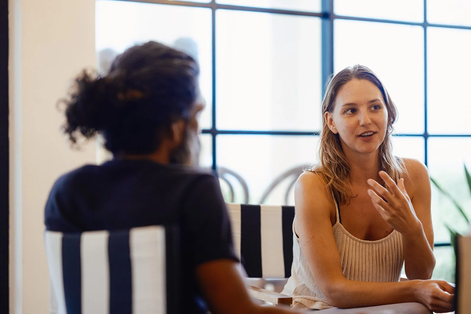 Two women having a conversation at a table in a bright room with large windows in the background.