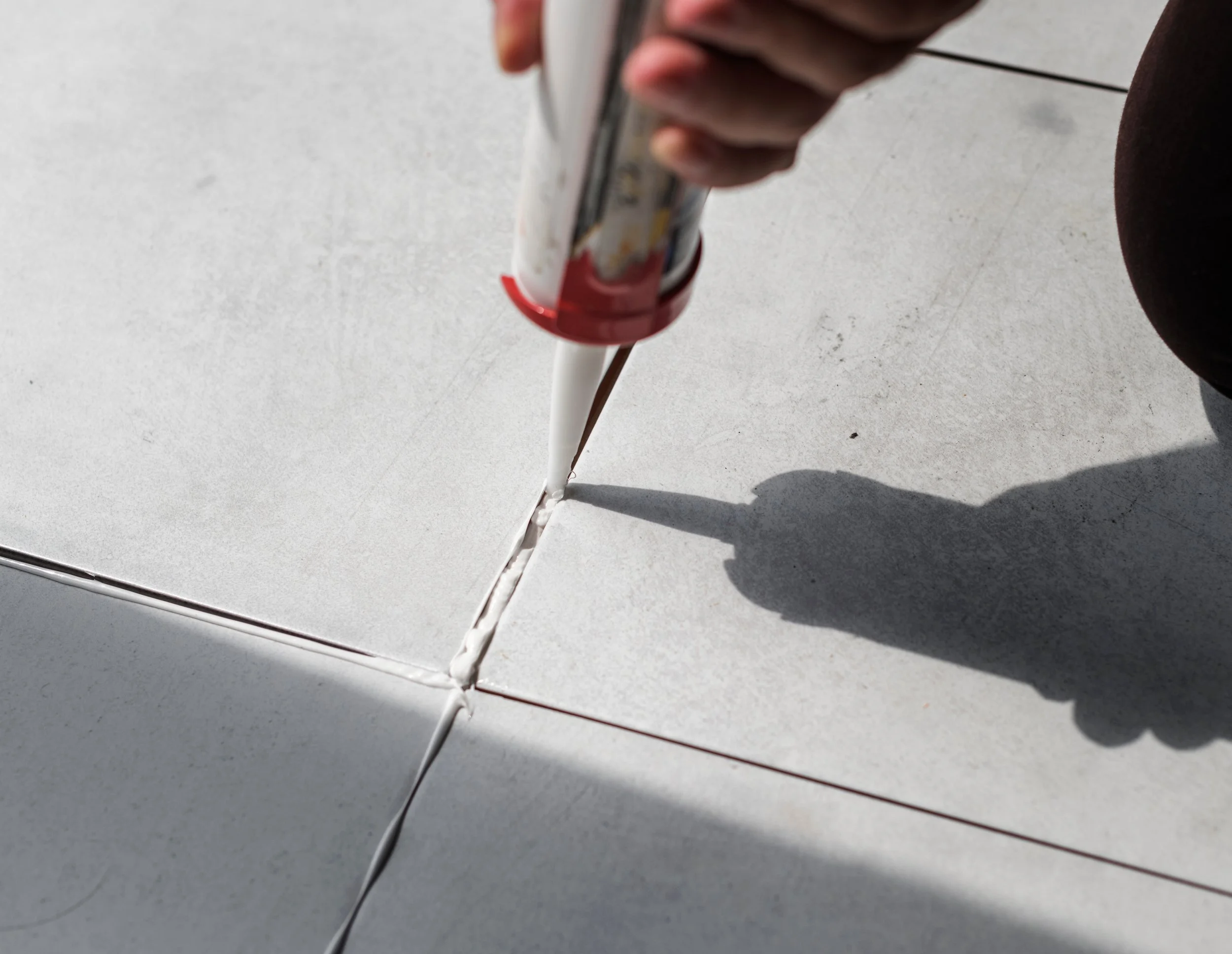 A hand holding a caulk gun while sealing tiled floors.