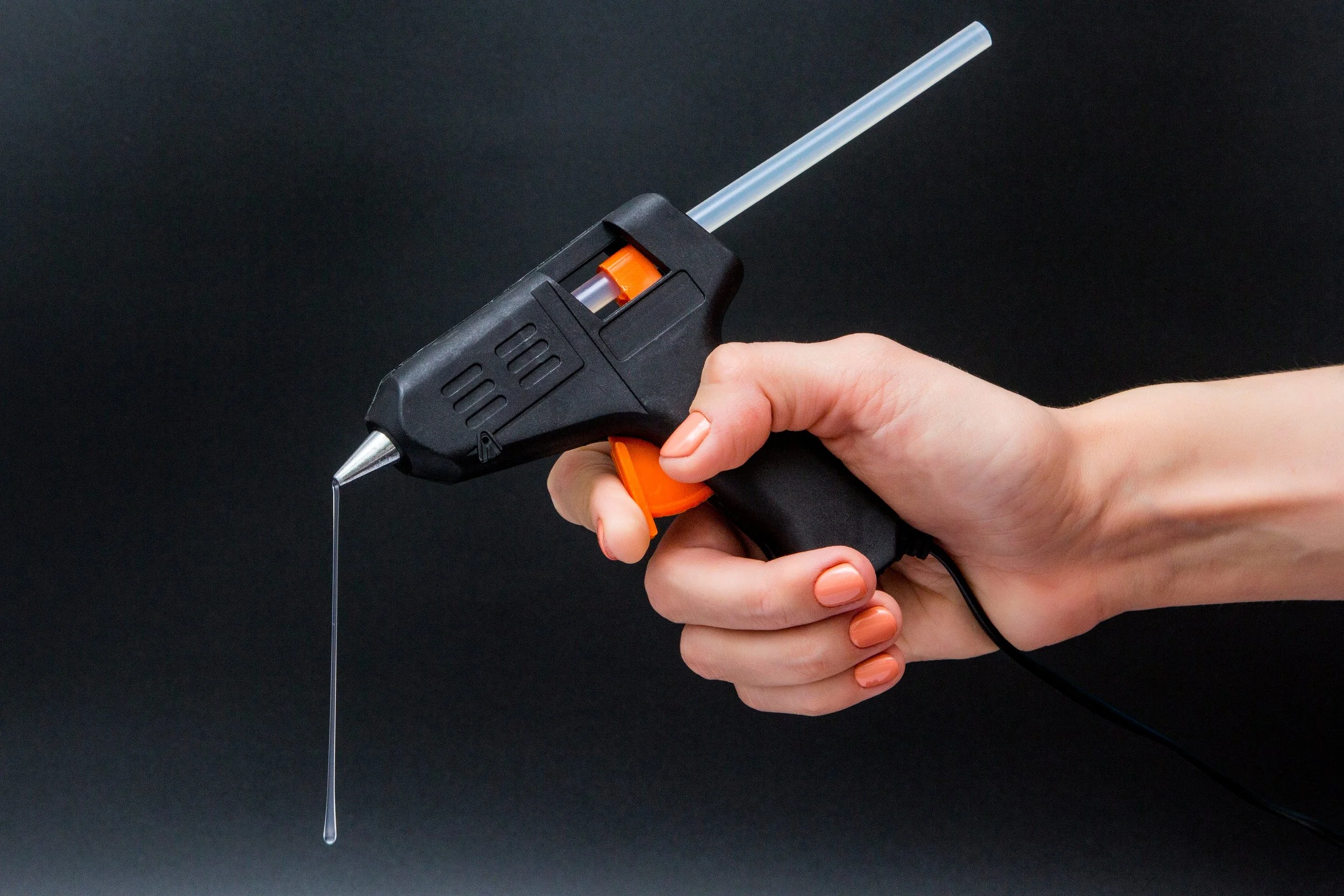 A hand holding a hot glue gun with glue dripping out against a dark background.