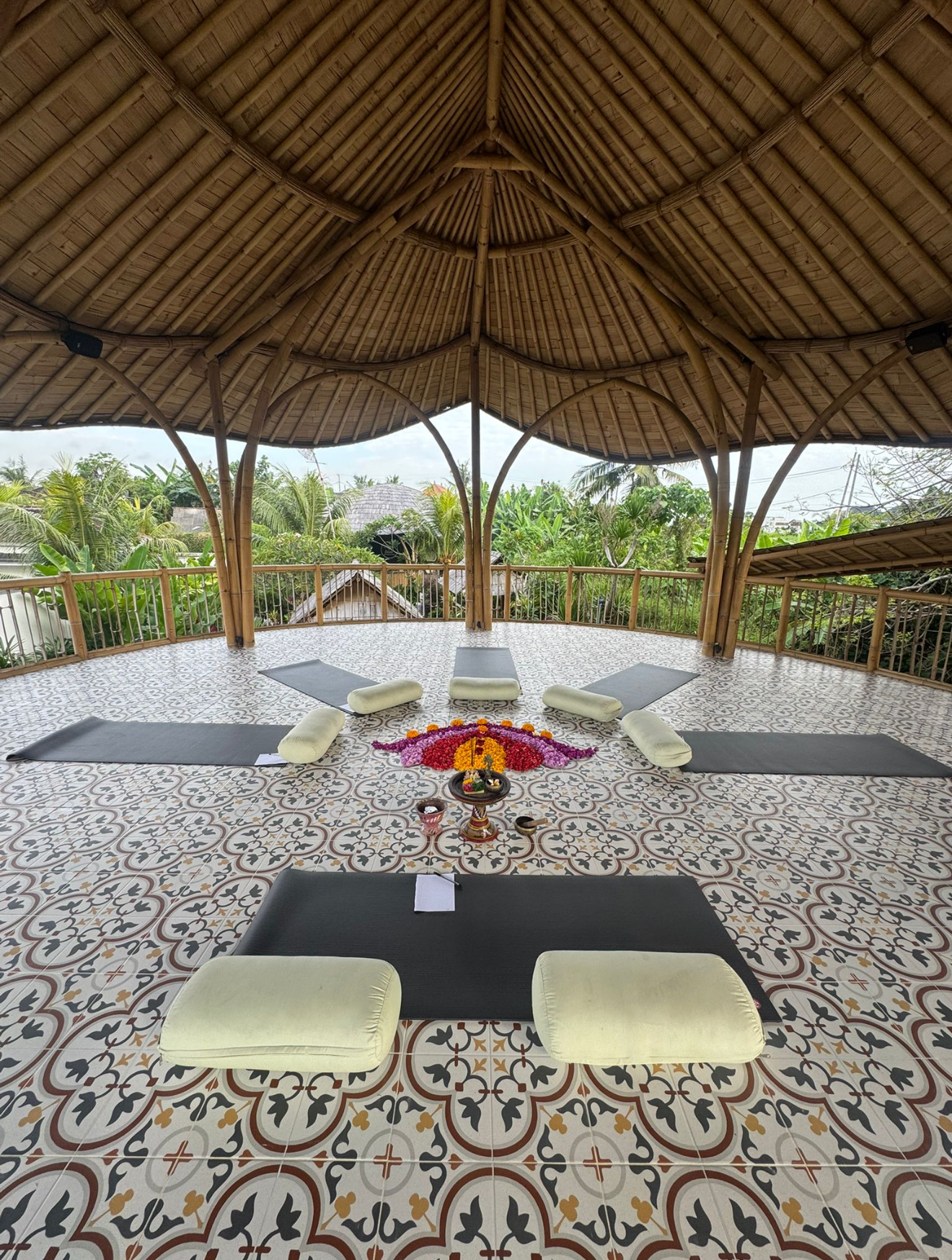 Open-air bamboo-roofed yoga or meditation space with mats, cushions, and floral decoration, overlooking lush greenery.