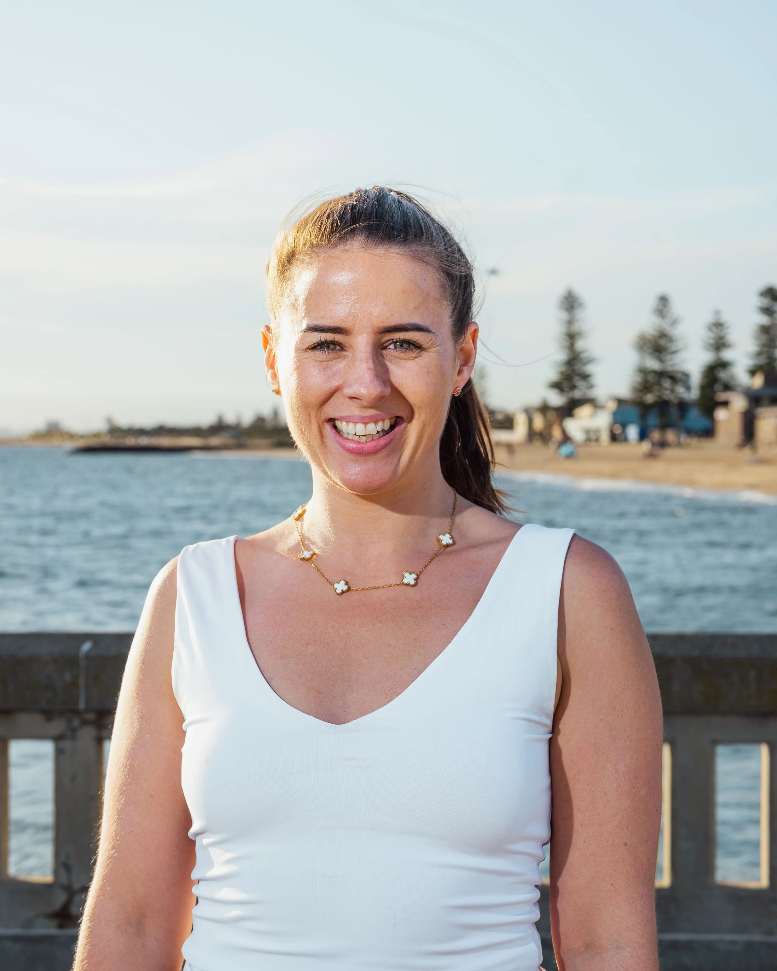A smiling woman with a ponytail smiling at the camera, standing outdoors by the water, with houses and trees in the background.