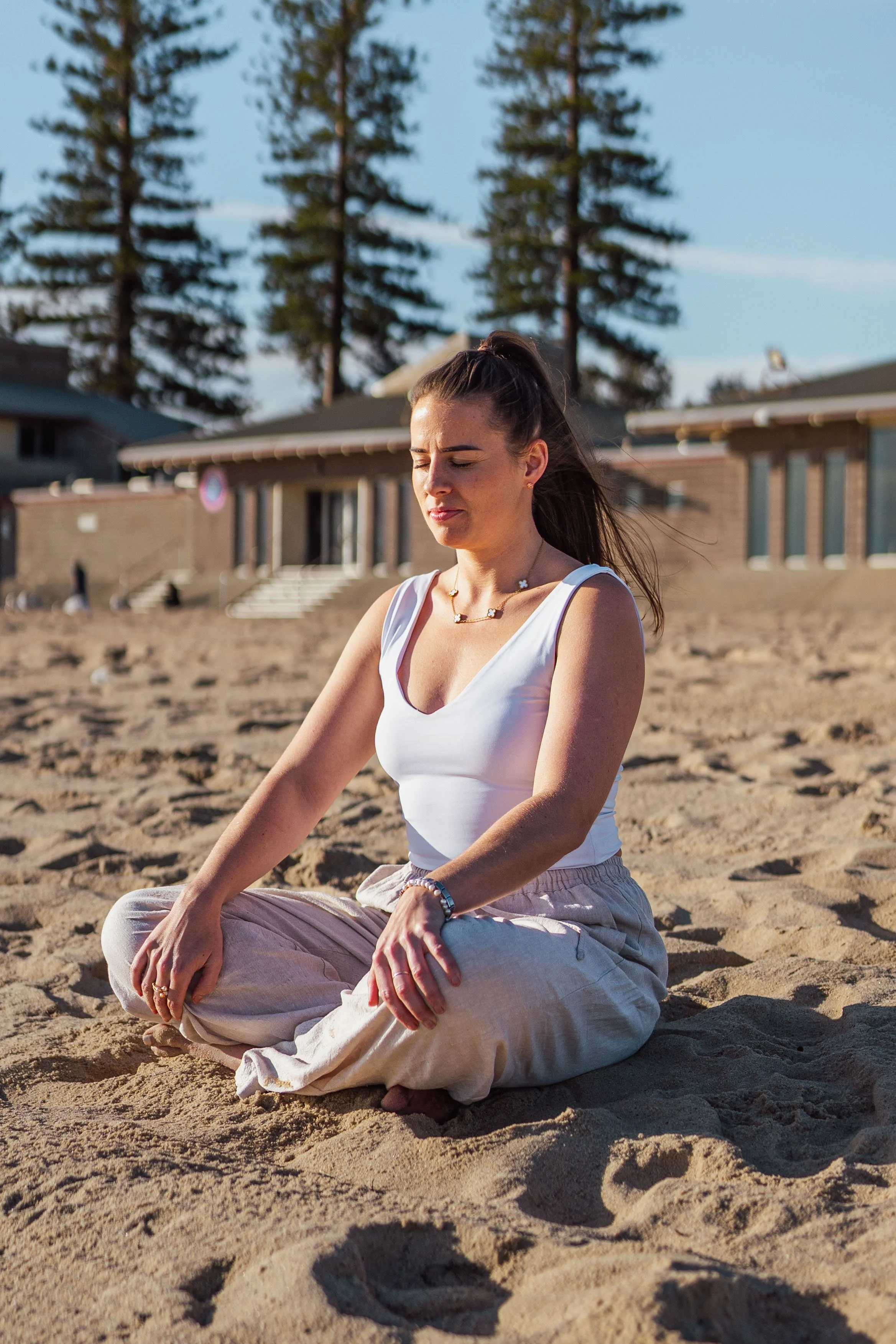 A woman practicing meditation on a sandy beach, seated cross-legged with her eyes closed, wearing a white tank top and loose pants, with houses and tall trees in the background.