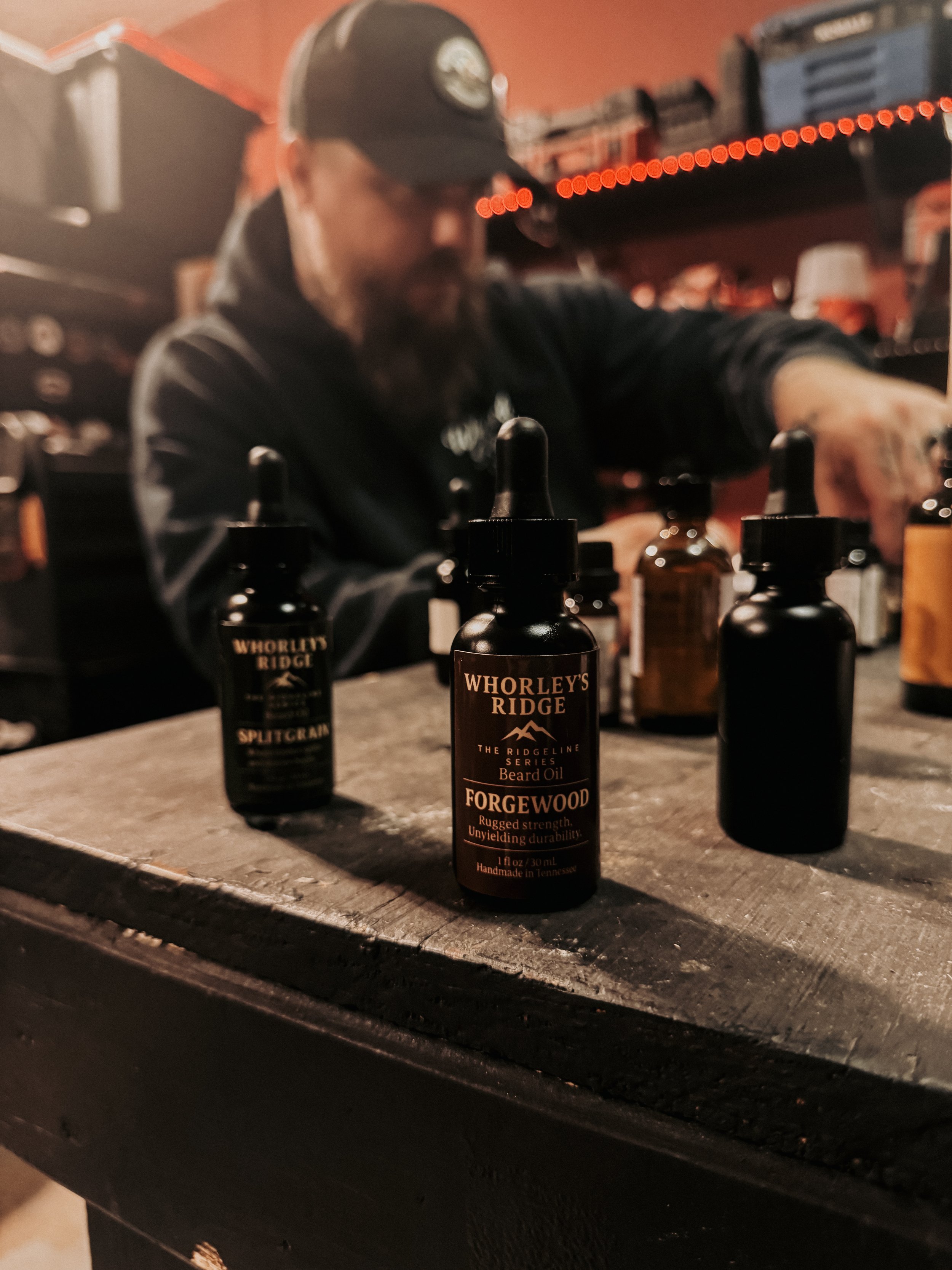 A man in a black cap and hoodie working with bottle dropper bottles of beard oil on a rustic wooden table, with a slightly blurred background of shelves and tools.