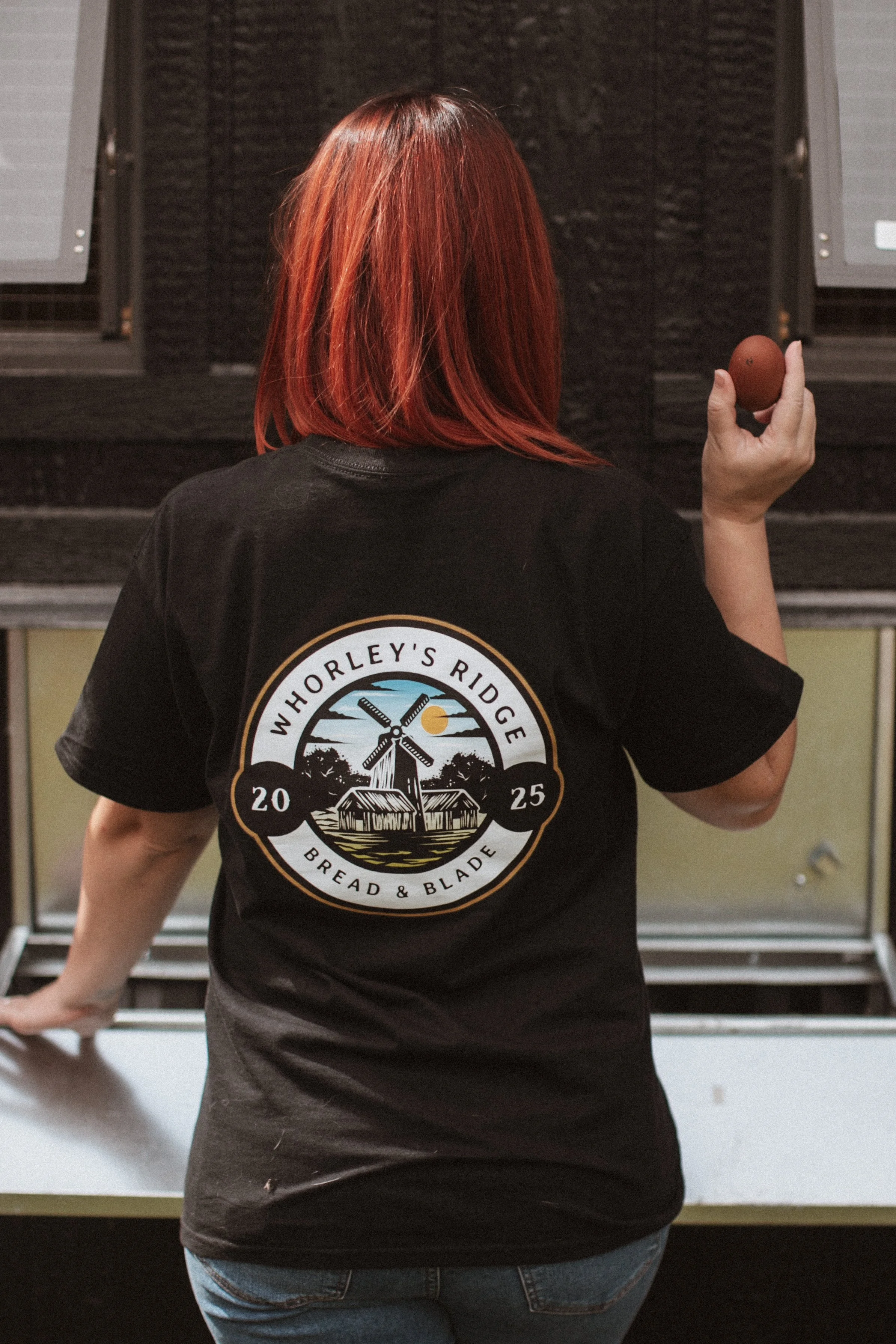 Person with red hair wearing a black shirt with a logo, holding an egg, standing in front of an oven or bakery equipment.