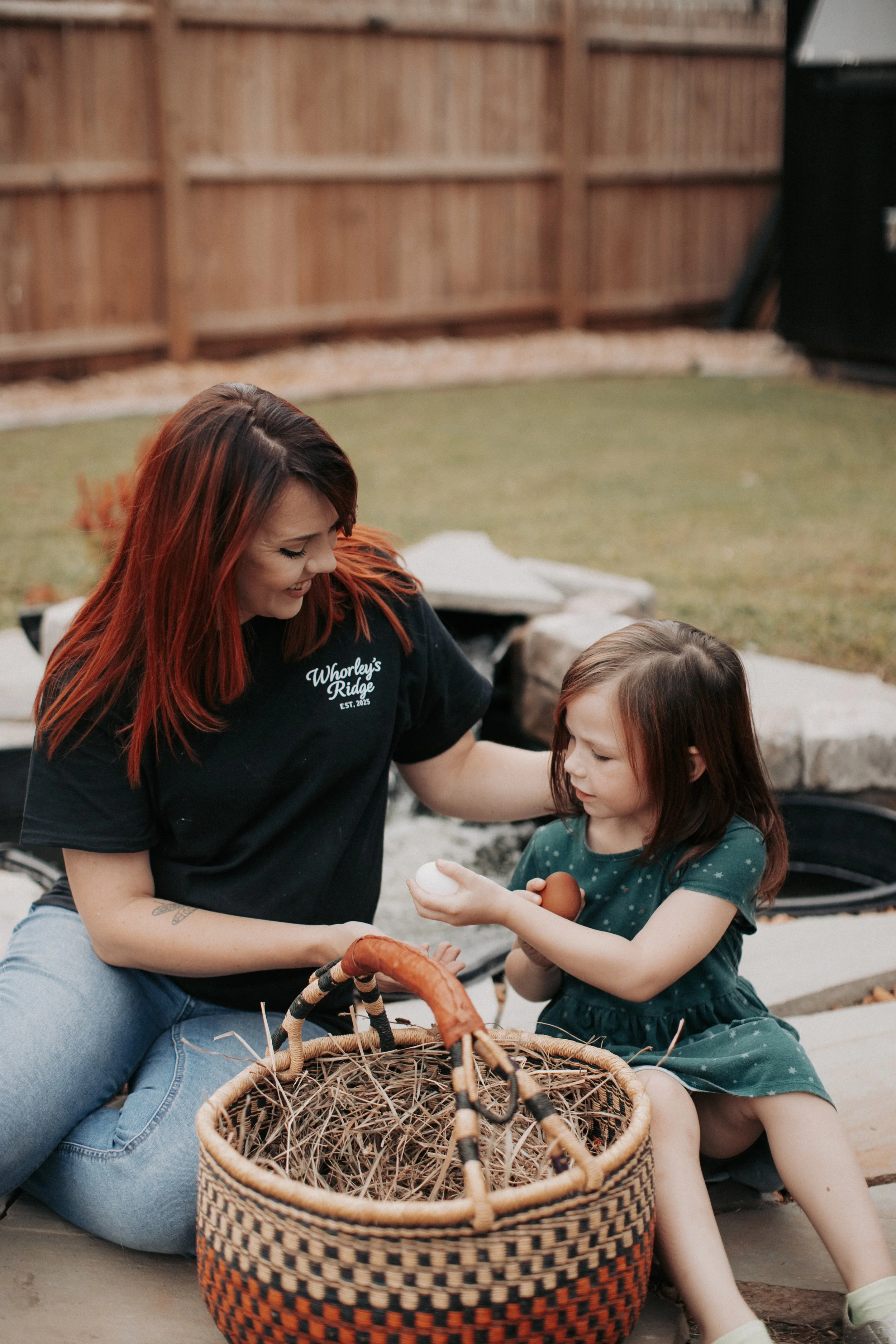 A woman and a young girl sitting outdoors near a fire pit, sharing a basket of eggs and holding one egg each, smiling at each other.
